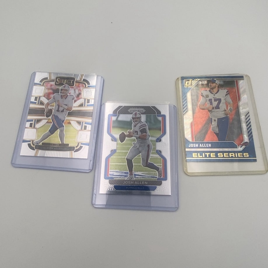 Josh Allen Trading Card Lot: This lot features three Josh Allen football trading cards from different sets, including Panini Select, Panini Prizm, and Donruss Elite Series. Each card is protected in a clear plastic sleeve. The