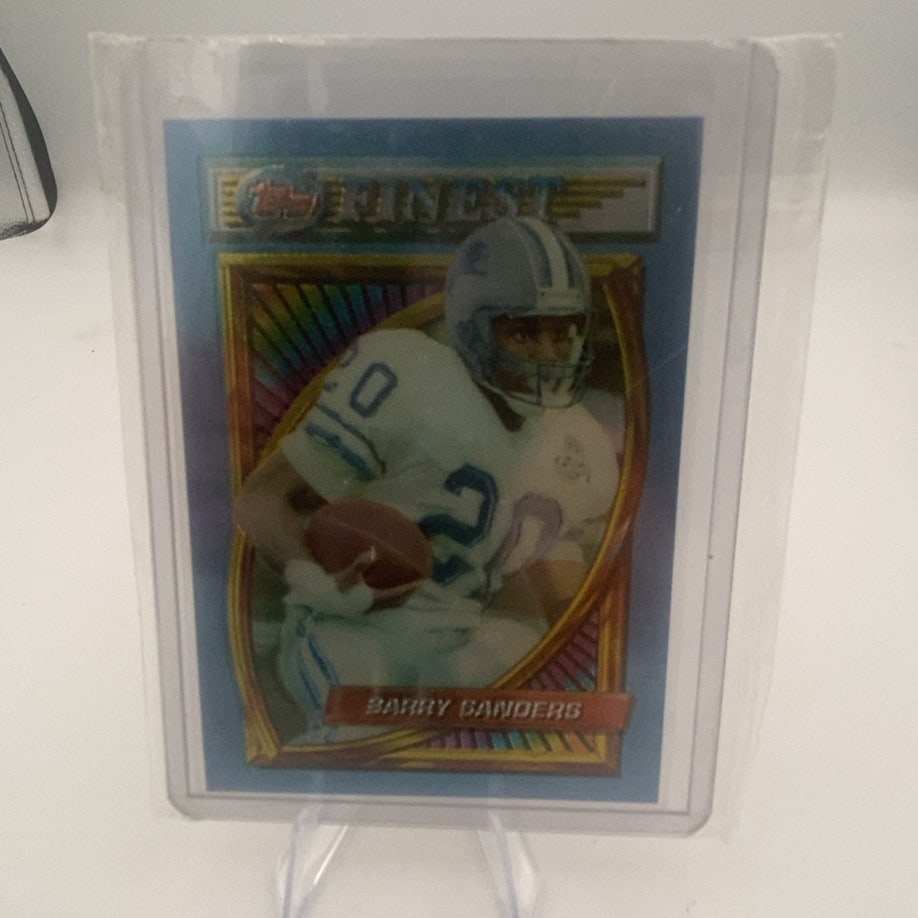 Topps Finest Barry Sanders: This is a Topps Finest trading card featuring football player Barry Sanders. The card is encased in a protective plastic sleeve and displays a holographic border design common to the Finest series.