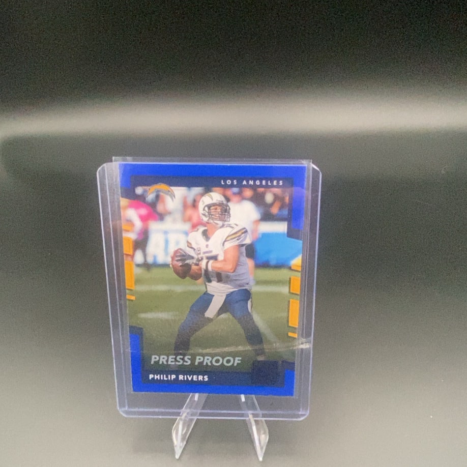 Philip Rivers Press Proof Card: This is a collectible trading card featuring Philip Rivers of the Los Angeles Chargers. The card is labeled as a 'Press Proof' version and is housed in a protective plastic sleeve.