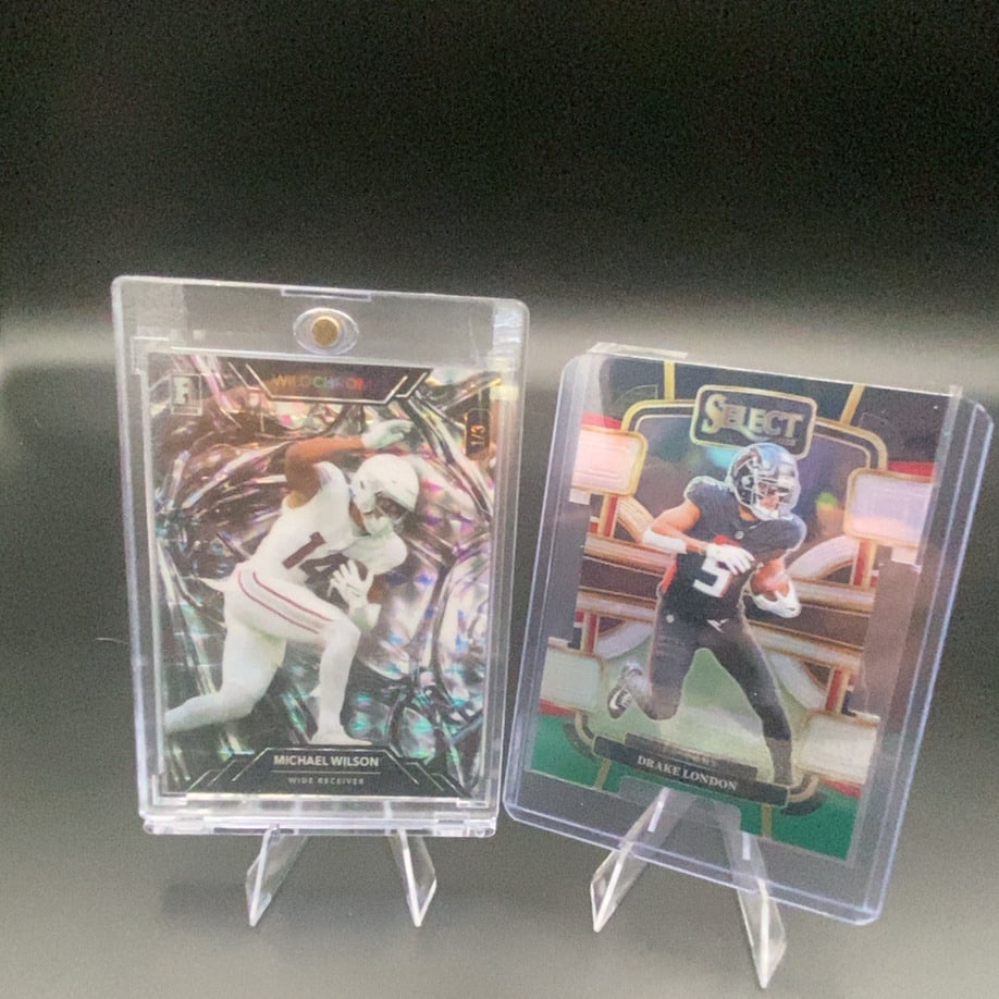 2023 Wild Card Michael Wilson and 2022 Panini Select Drake L: This image features two football trading cards protected in plastic cases. One card depicts wide receiver Michael Wilson from the 2023 Wild Card set, while the other shows wide receiver Drake London f