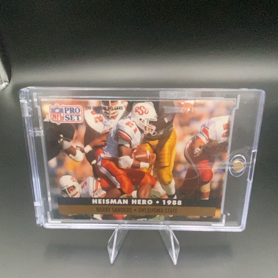 1988 Pro Set Barry Sanders: This is a 1988 Pro Set Heisman Hero trading card featuring Barry Sanders of Oklahoma State. The card highlights his 1988 collegiate season and is protected in a clear magnetic holder.