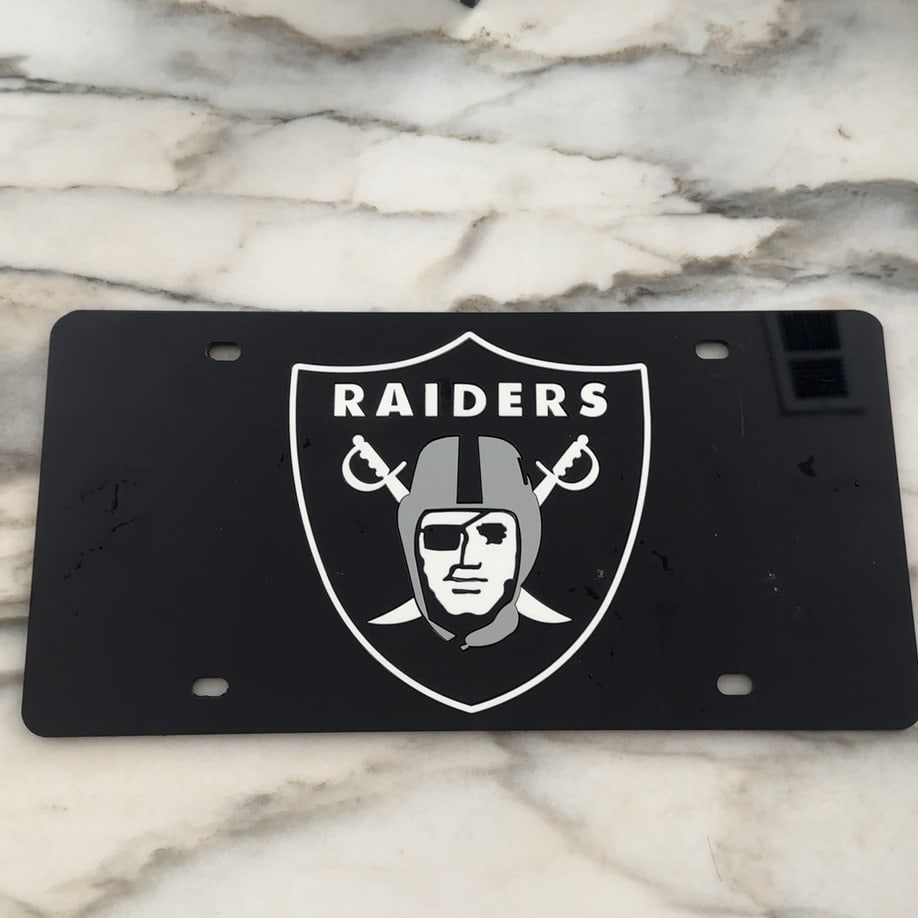 Las Vegas Raiders License Plate: This is a decorative license plate featuring the classic logo of the Las Vegas Raiders NFL team. The design showcases the iconic shield and pirate mascot on a black background.