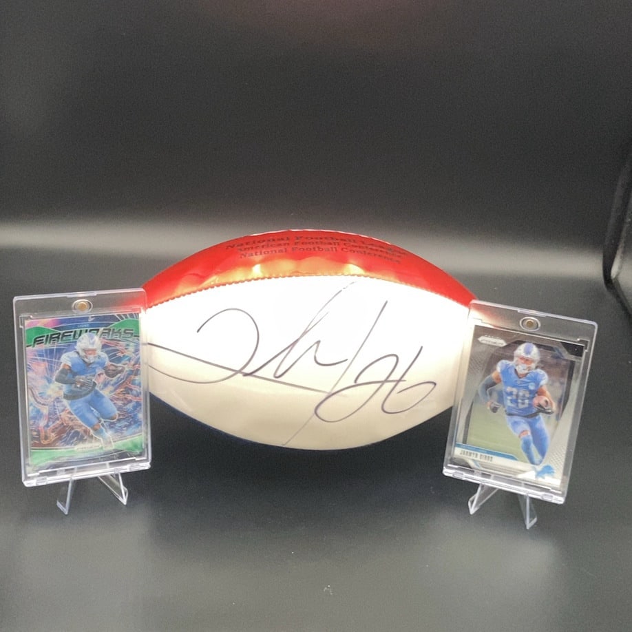 Jahmyr Gibbs Autographed Football and Trading Cards: lot features a football autographed by Detroit Lions running back Jahmyr Gibbs. It is displayed alongside two Prizm brand trading cards featuring Jahmyr Gibbs, one of which is part of the Fireworks in