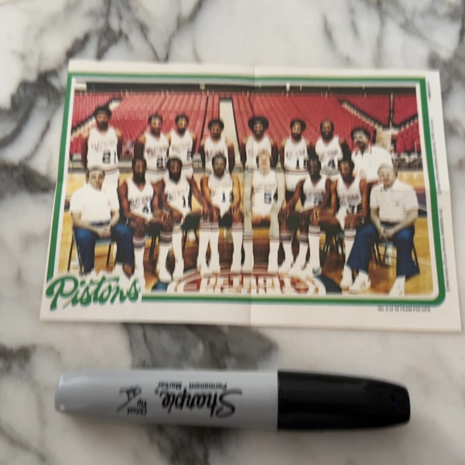 1980s Detroit Pistons Team Photo: A vintage team photograph of the Detroit Pistons basketball team. The image shows the players and coaching staff in their uniforms on a court. It is marked as part of a team pin-up set.