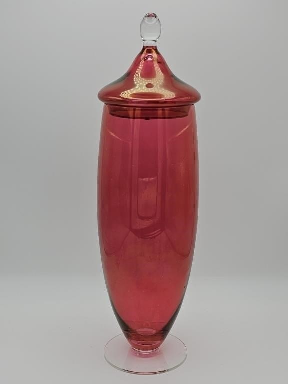 Red Glass Apothecary Jar: This is a tall, elegant apothecary jar or canister crafted from vibrant red glass. It features a matching glass lid with a clear finial knob on top. The piece sits on a simple, clear circular base.