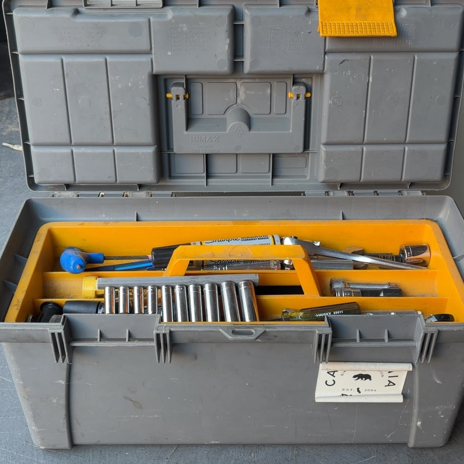 Rimax Tool Box: This is a grey and yellow Rimax brand tool box. It features an open top with various hand tools and a socket set inside. The box has a label on the front that displays a bear icon and the text EST.