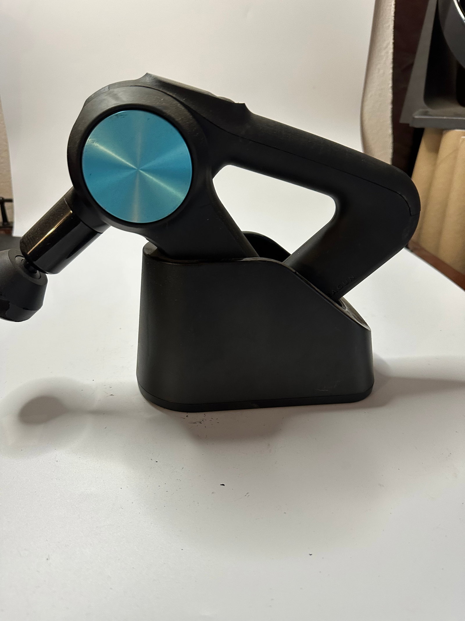 Theragun Percussive Therapy Device: This is a black Theragun percussive therapy massage device shown with its charging base. The unit features a distinct triangular handle design and a prominent blue circular accent on the side casing.