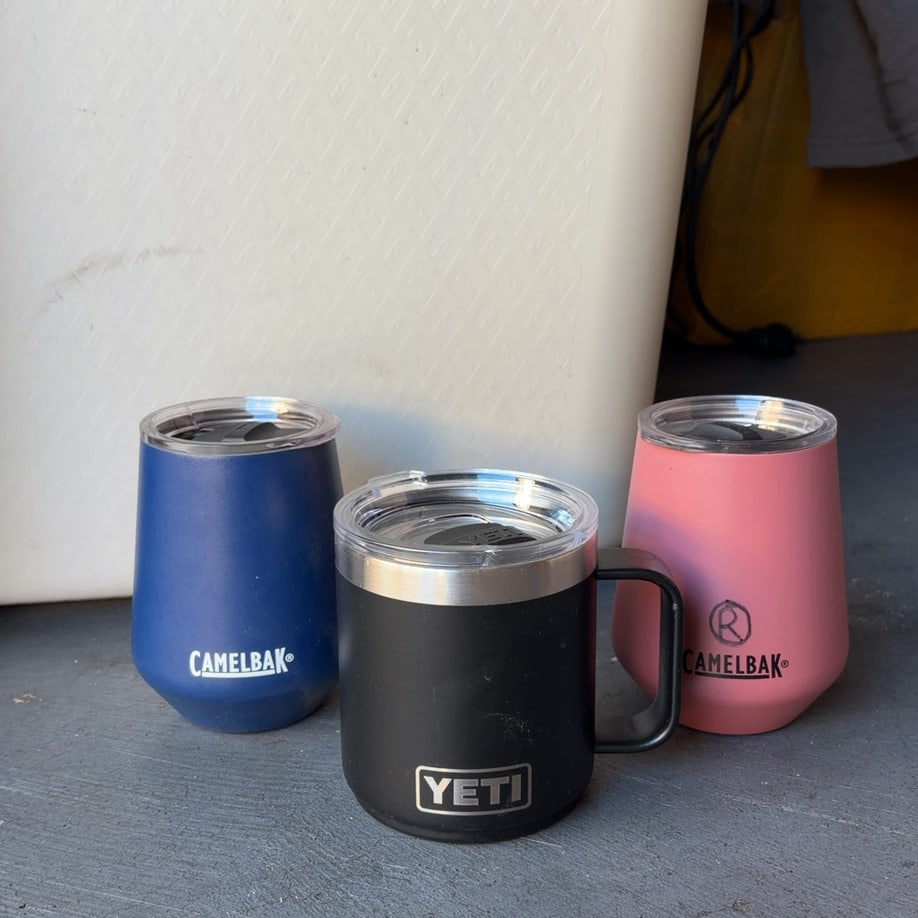CamelBak and YETI Insulated Mugs: This lot features three insulated drinkware items, including two CamelBak mugs in blue and pink, and one black YETI handled mug. Each mug comes with a clear lid and features the respective brand's
