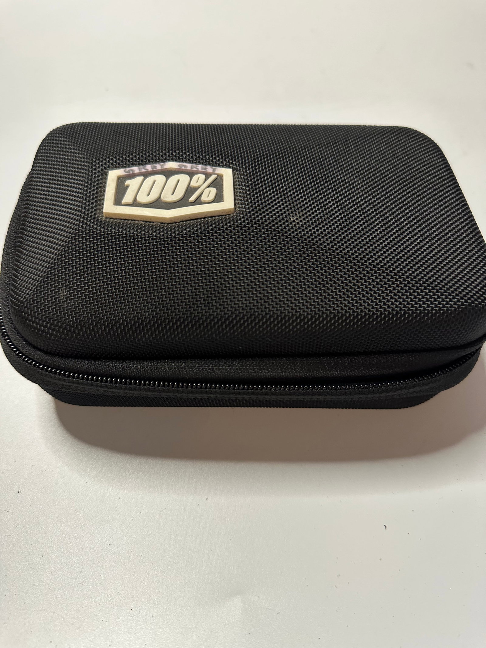 100% Eyewear Protective Case: This is a black, textured hard-shell storage case featuring the 100% branding logo on the front. It is designed to provide protective storage for eyewear and related accessories.