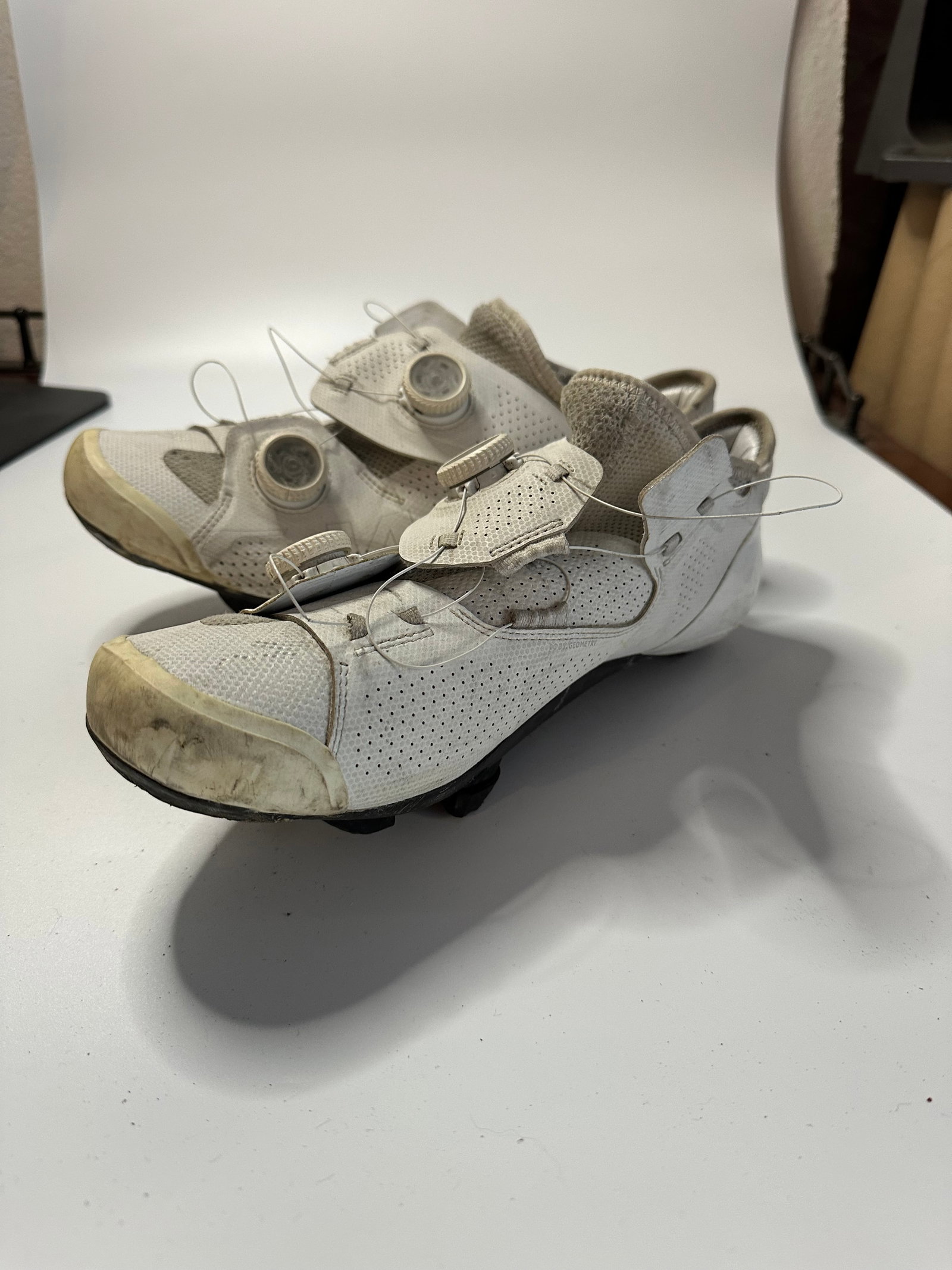 White Cycling Shoes: A pair of white cycling shoes featuring a unique BOA-style lacing system. The shoes have a perforated upper design and show signs of wear on the front toe areas.