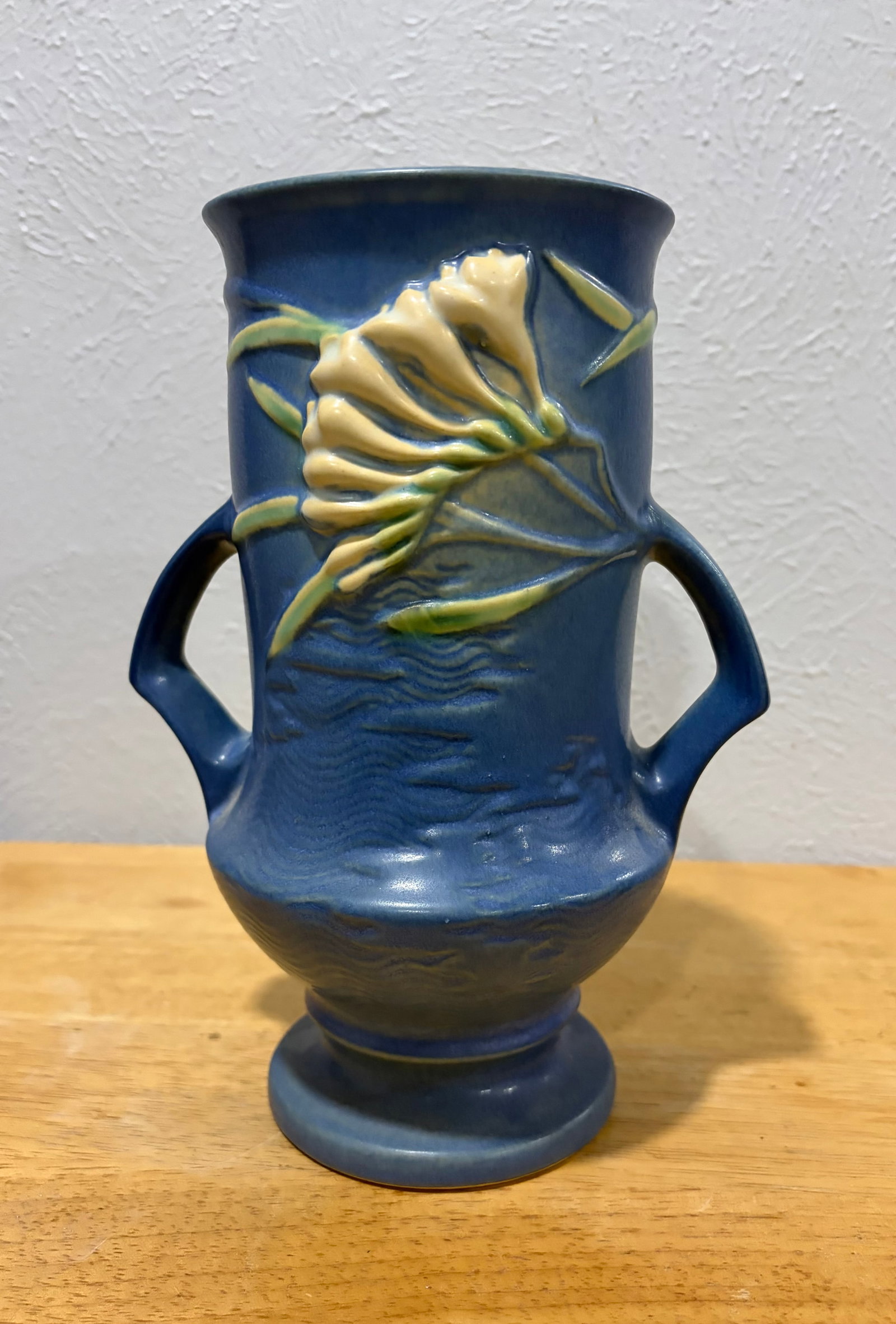 Freesia Roseville Pottery Ceramic Vase: A stunning Roseville pottery vase featuring the Freesia design. This elegant piece showcases a rich blue background with raised floral motifs in soft yellow and cream tones, accentuated by vibrant gre