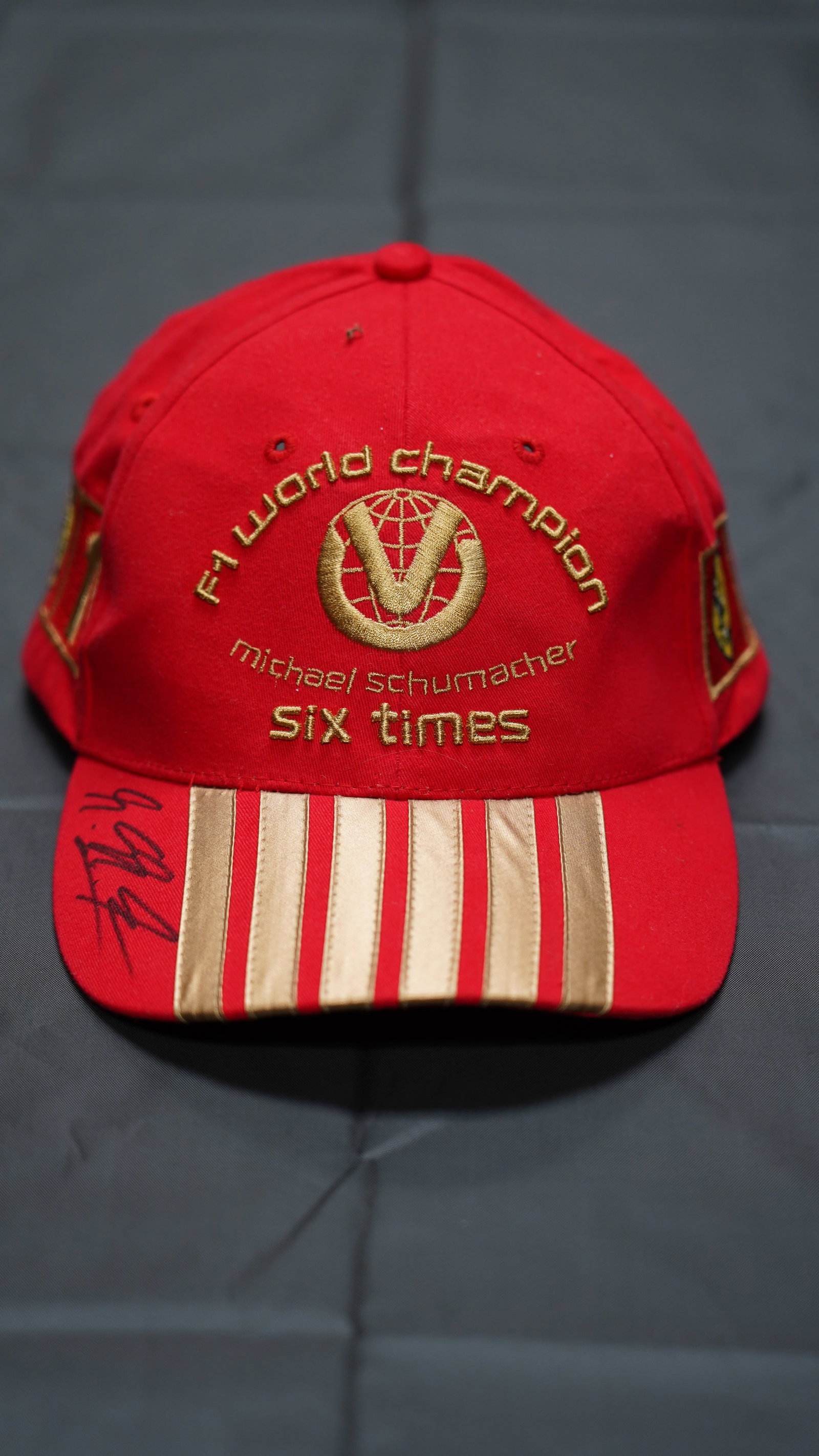 Michael Schumacher Signed Ferrari F1 “Six Times World Champion” Hat: Official Ferrari Michael Schumacher Collection cap celebrating his six World Championships. Features premium gold embroidery, Ferrari shield patch, and #1 driver insignia.Hand-signed autograph on the 