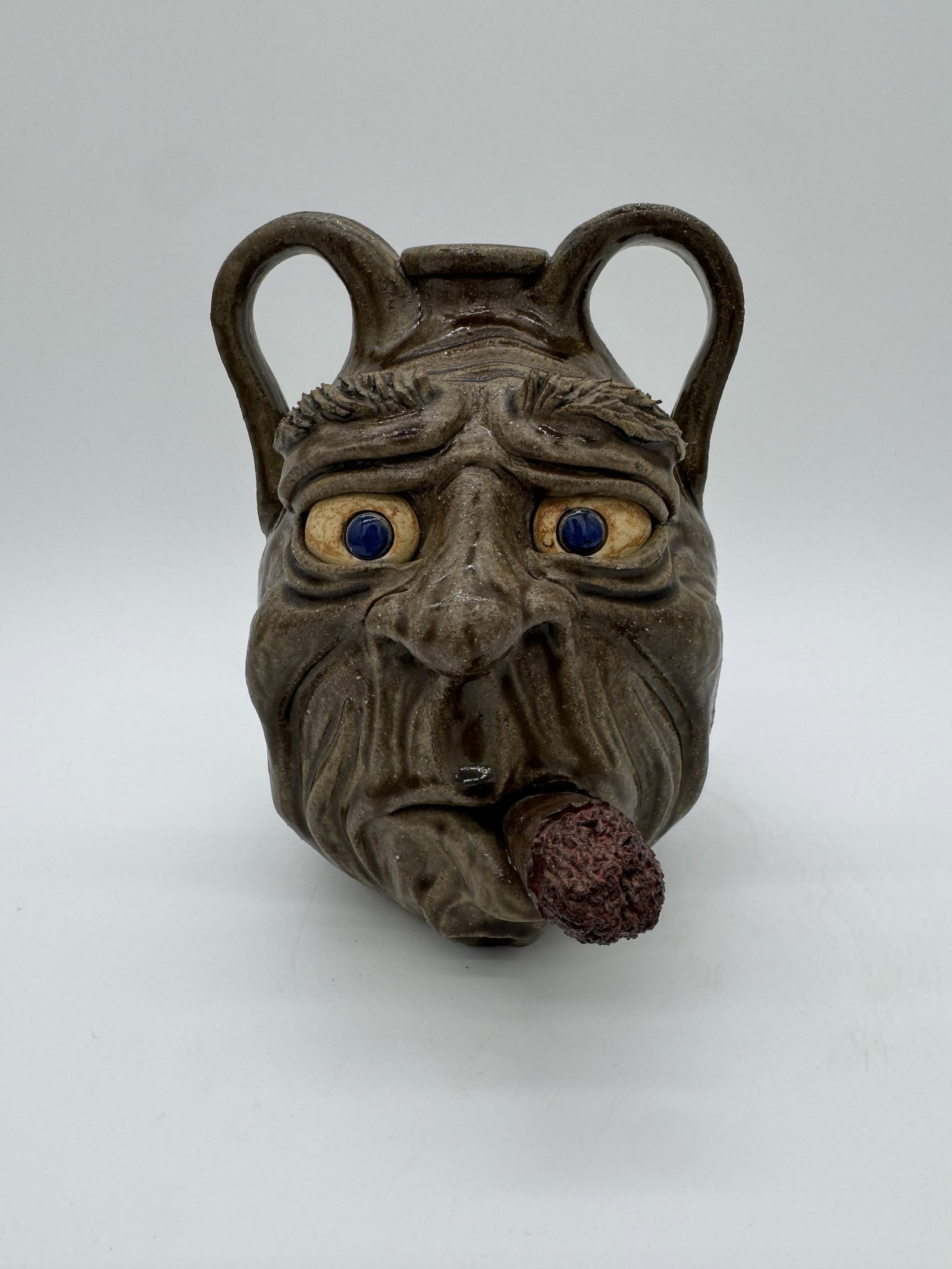 Turkey Merck Double Face Jug: A double face jug by Turkey Merck, featuring two contrasting expressions—one side depicted smoking a cigar, while the other shows a lazy eye. This example includes blue eyes that add contrast and ch