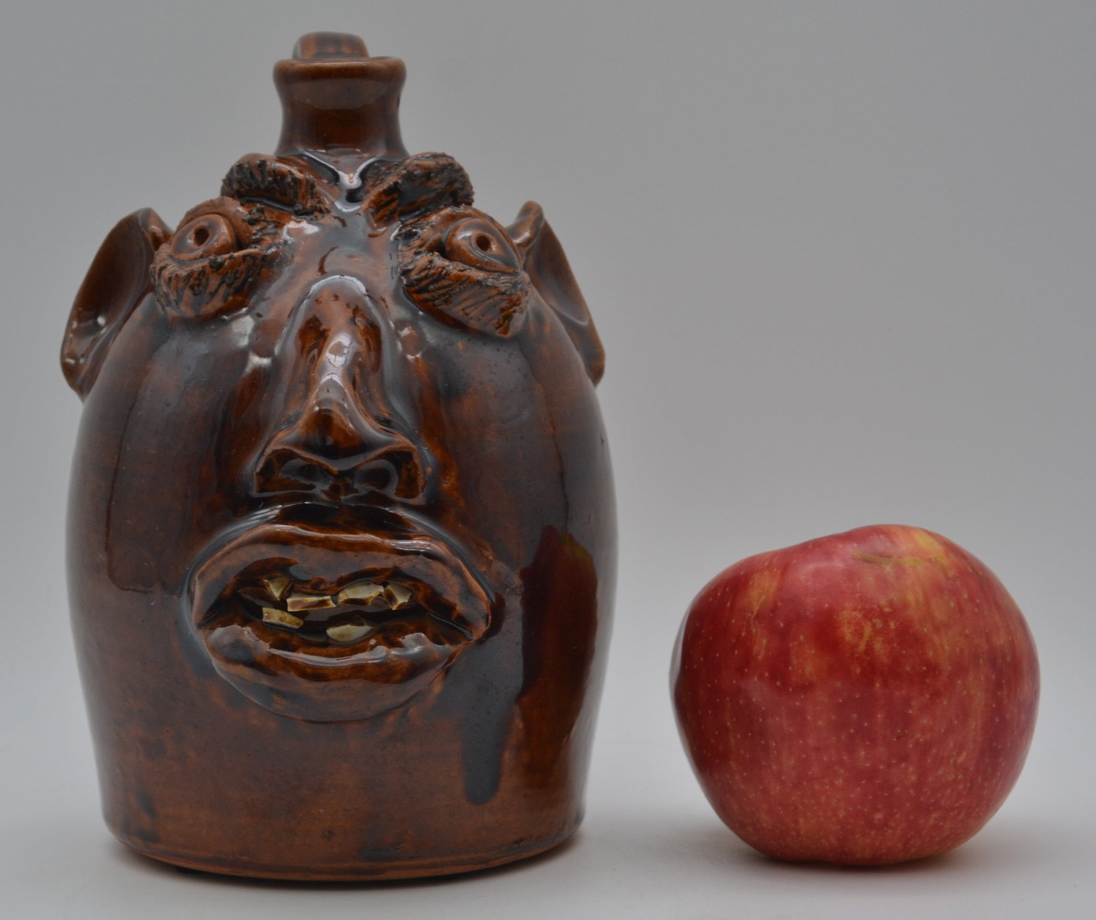 Robert Brown Face Jug: A face jug by Robert Brown featuring china plate teeth and a deep, rich brown glaze. This striking example has excellent character, strong craftsmanship, and beautiful surface throughout. Signed on th