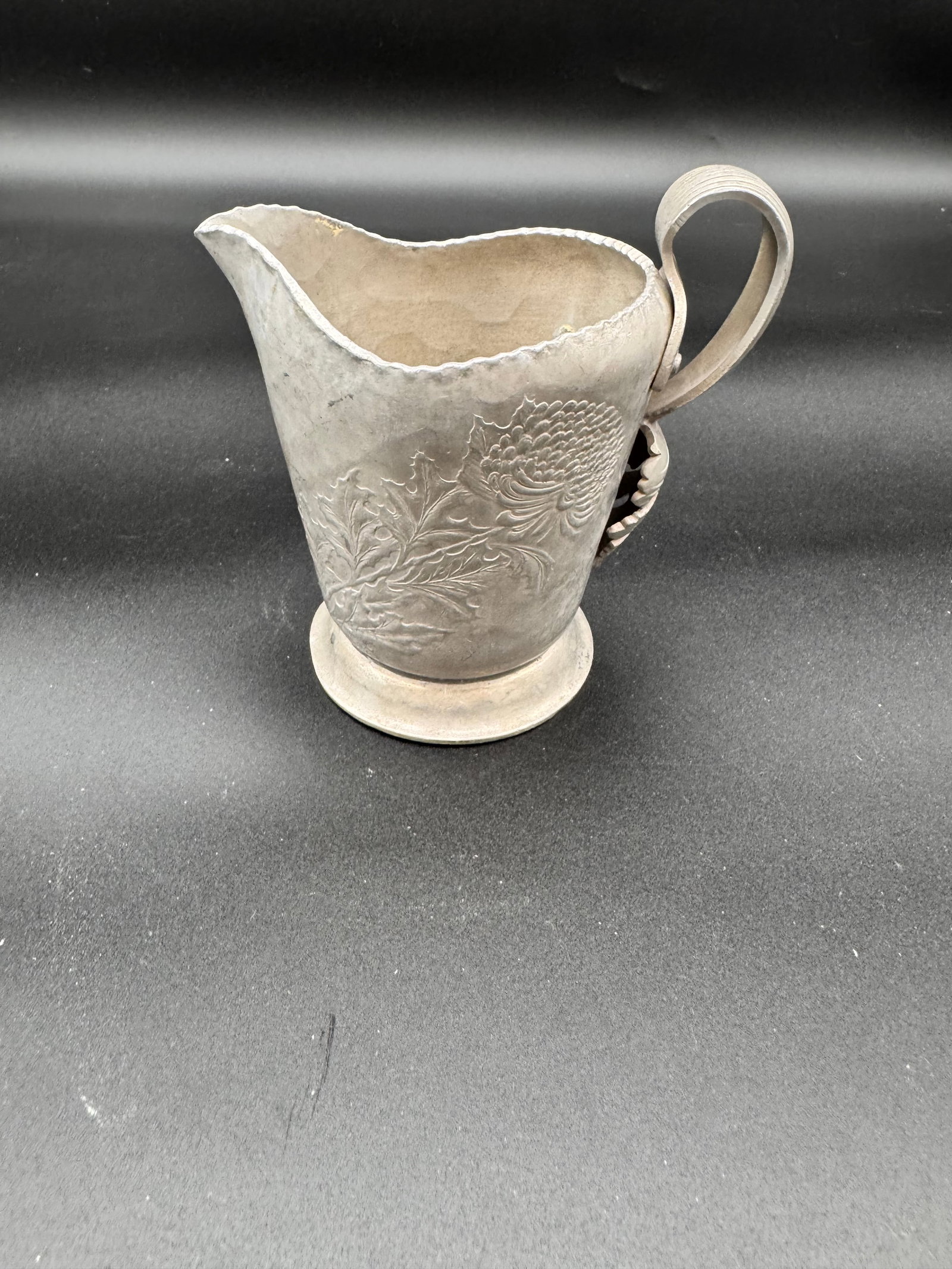 Pewter Pitcher: This mid century pitcher has a floral design with a cured handle and a pouring spout. The surface has a matte, neutral toned finish.