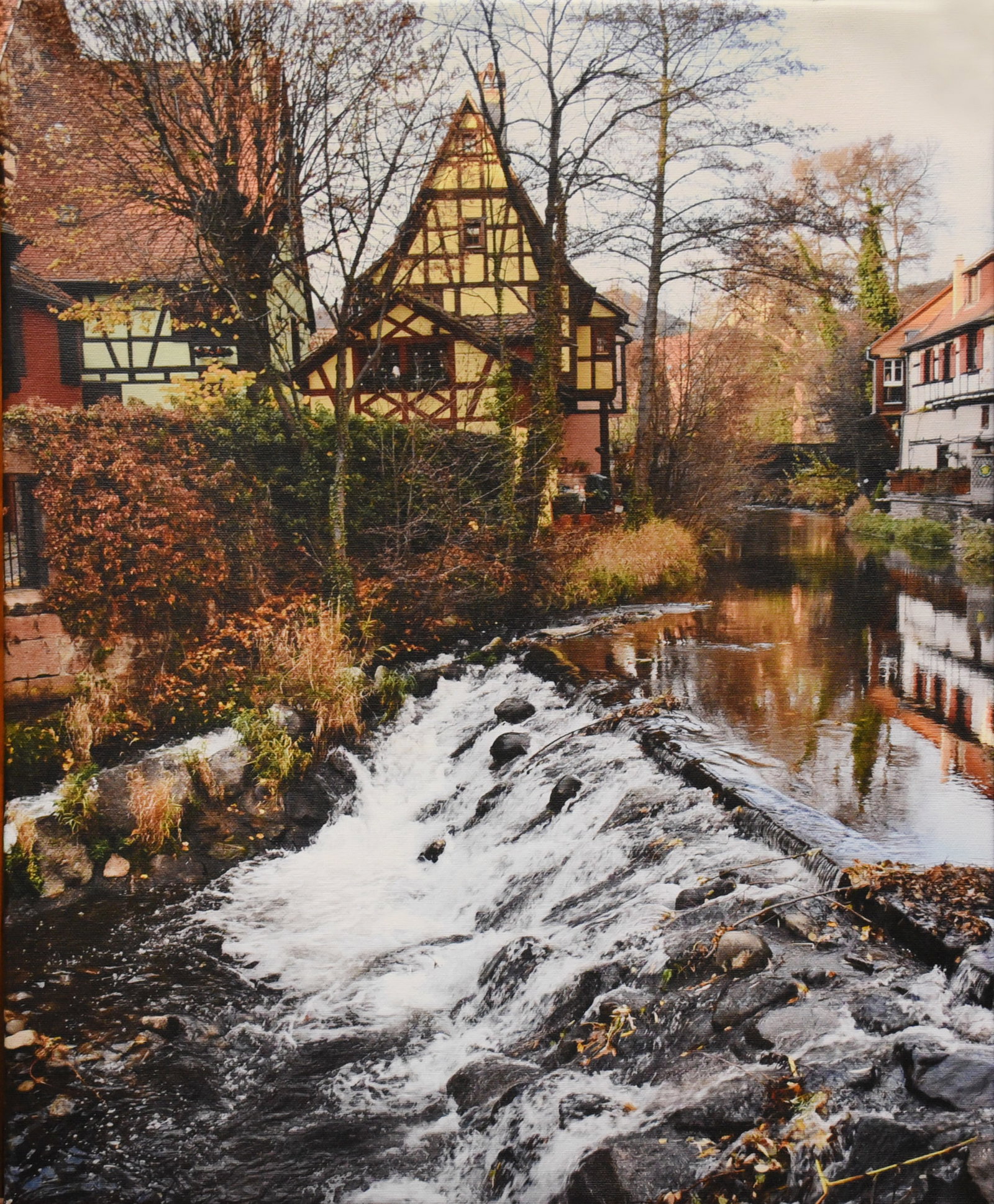 Village on the Water: Bring the charm of the French countryside into your home with 'Alsatian Charm,' a beautifully captured scene printed on canvas. This 16" x 20" piece depicts a quaint village nestled along a gently