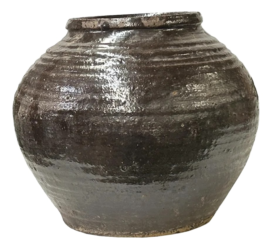 Vintage Earthy Rustic Brown Black Village Rural Pottery Ceramic Pot Vase: The is a stout ceramic jar with a rounded body that tapers towards a narrow, slightly everted rim. The upper portion of the jar is covered in a glossy, dark rustic brown black glaze with subtle variat
