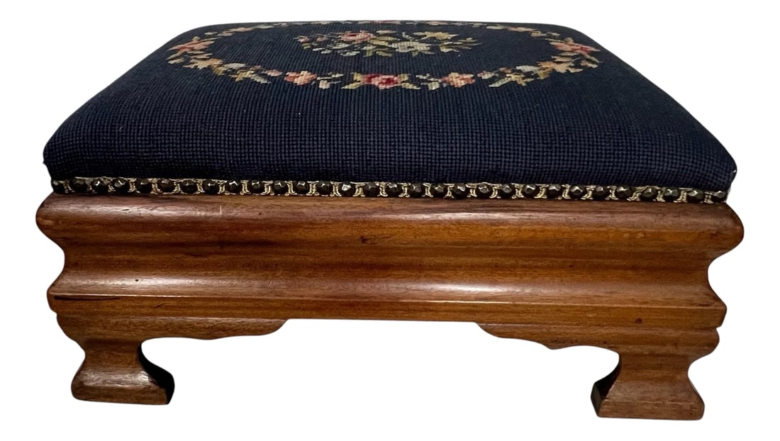 Antique Needlepoint Footstool: Pink and green roses on a navy background form a wreath around a larger bouquet of the same all on a antique mahogany base either a studded band separating between. Measures 19" w x 14" d x 9.5" h.