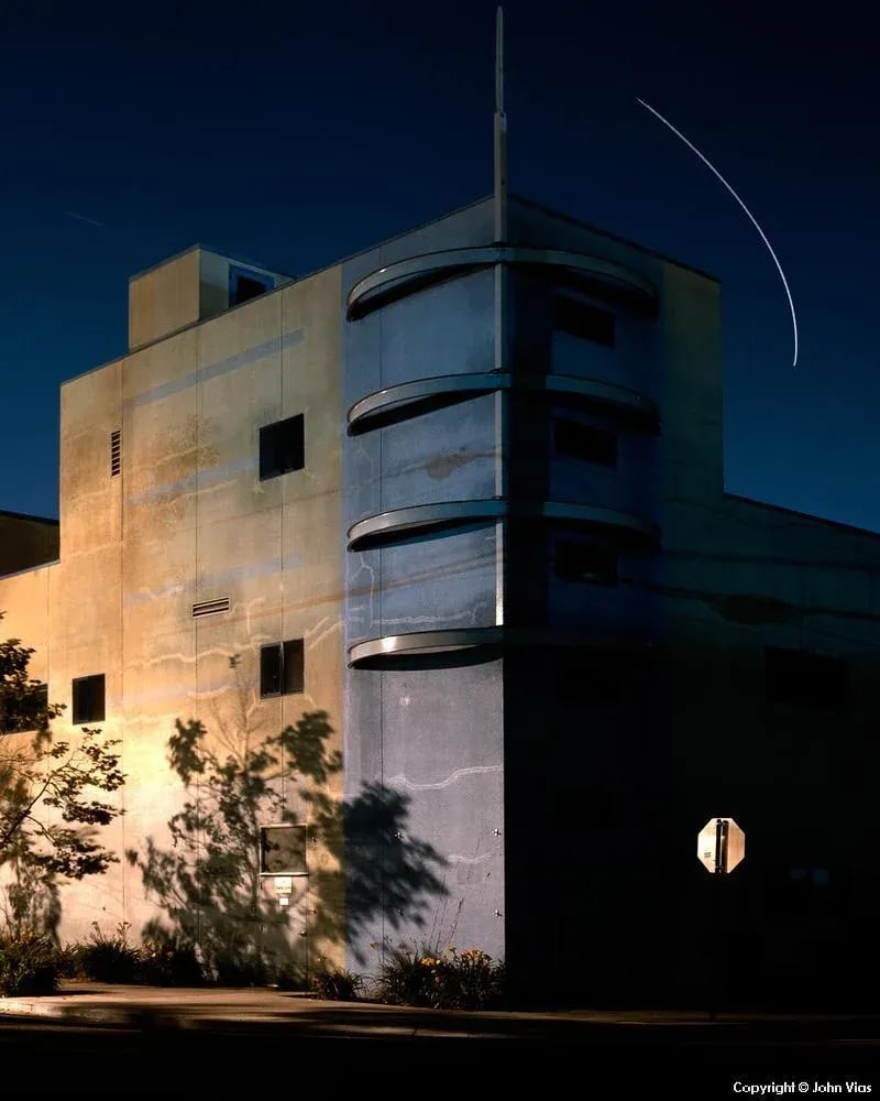 Contemporary Night Photograph "Banded Building" by John Vias: Note from the artist, John Vias: "Headlights, streetlights, and airplane trails can ruin an image. But in this one, the airplane trail adds to the result by echoing the building's bands." This 14" x