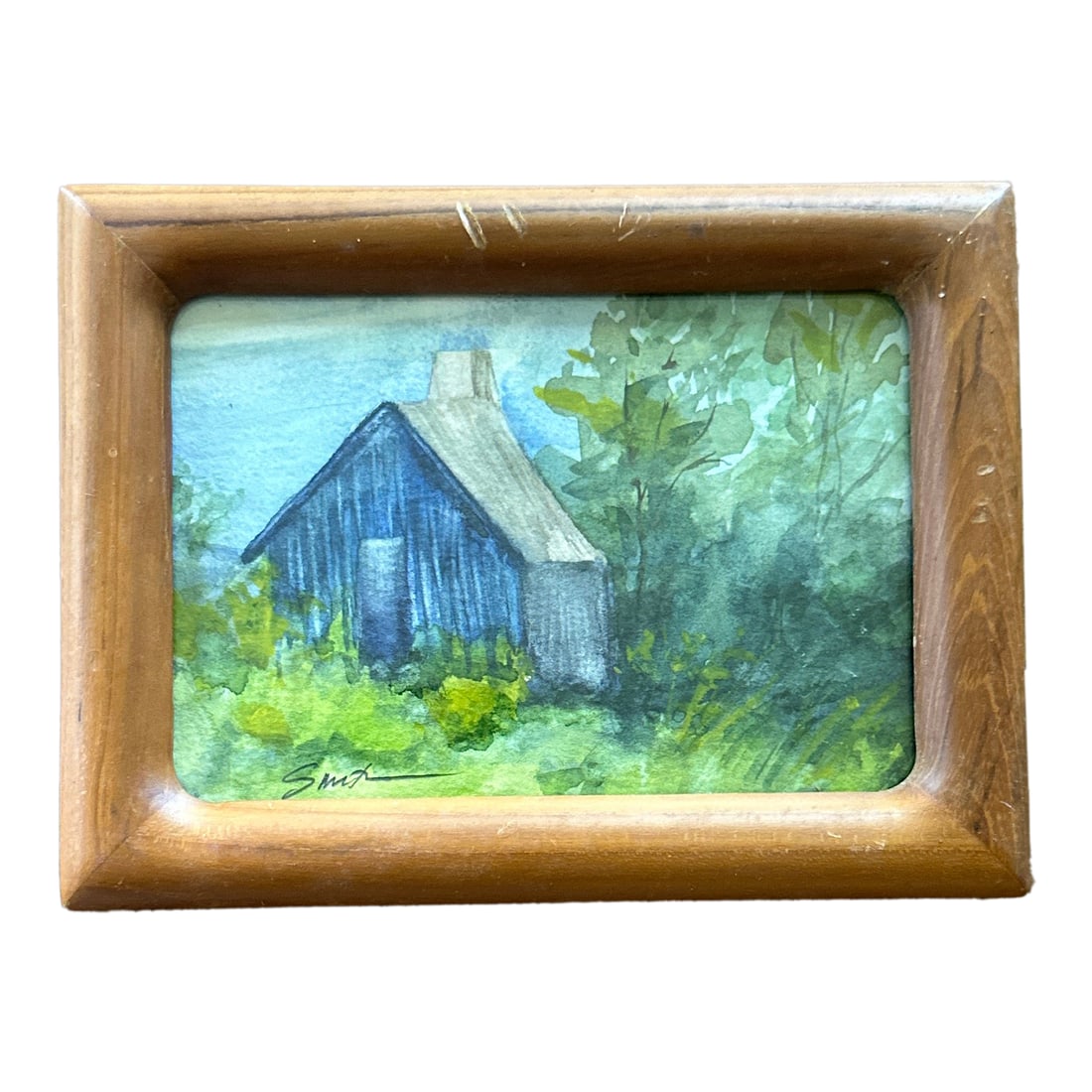 "Cabin in the Green" Framed Signed Original Watercolor Painting by Nancy Smith: This charming little painting evokes the nostalgia of quiet retreats and simpler times. "Cabin in the Green" shows a rustic blue cabin nestled in lush, overgrown greenery, offering a peaceful escape