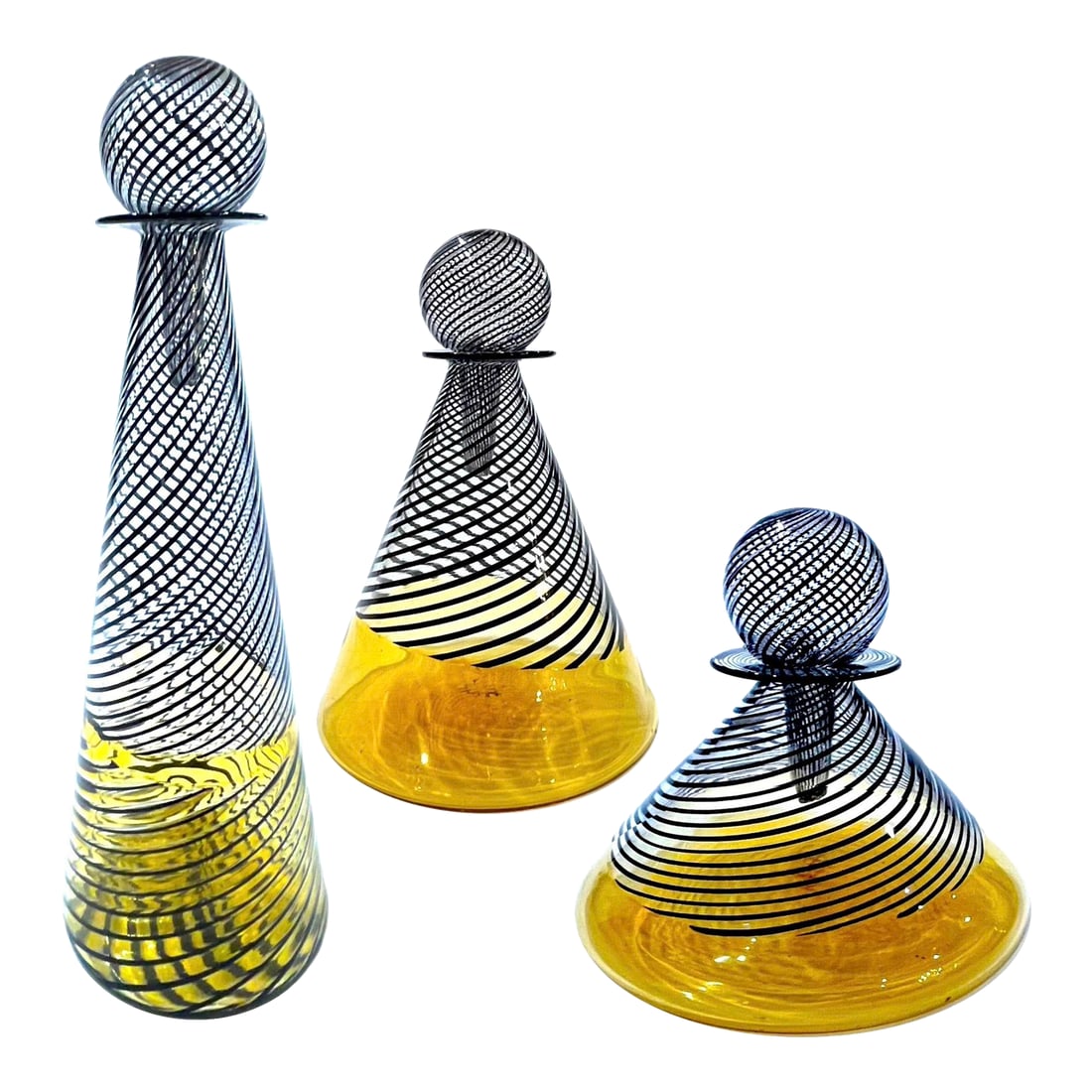 1960's Murano Attributed Latticino Glass Amber/Black Swirl Decanter Trio: A veritable tour de force of the glass blowers art. This set of three decanters with stoppers was blown using the latticino technique. Each piece features a swirl of black lines over a clear to amber 