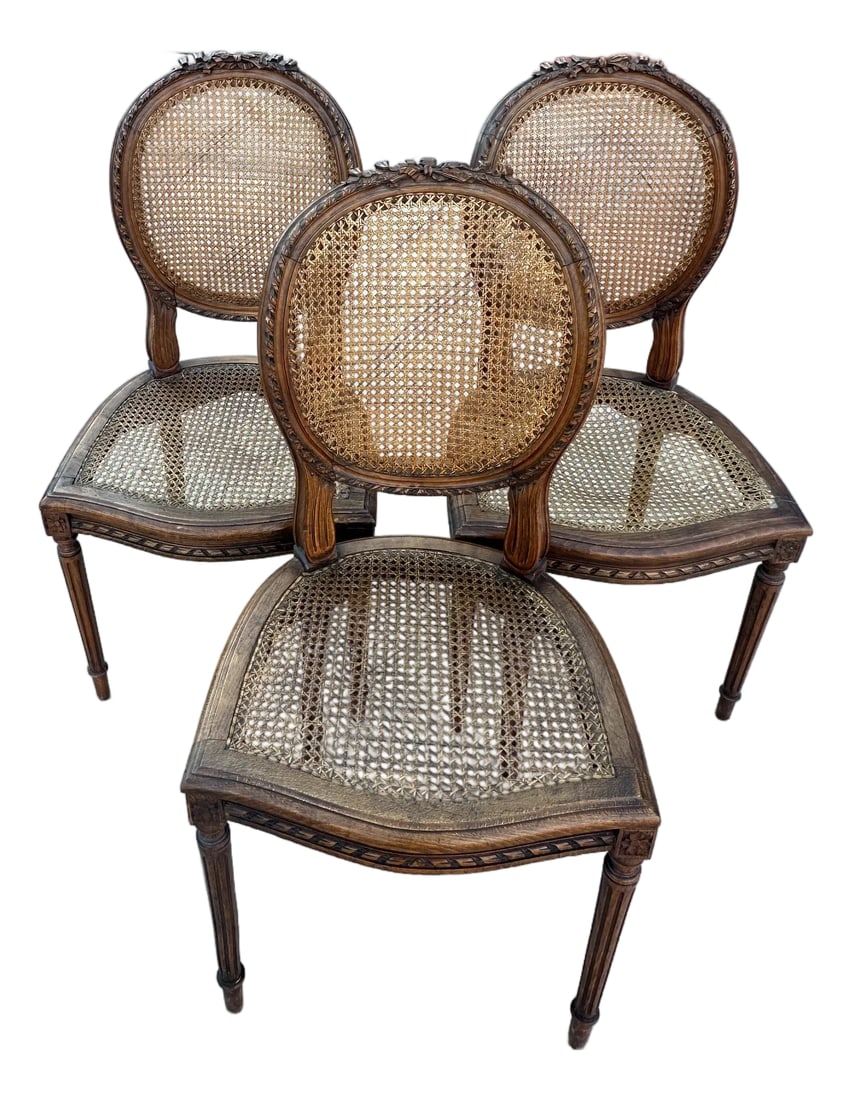 Vintage French Louis XVI Medallion Cane Back Chairs- A Set: Vintage French Louis XVI Style Chairs featuring carved walnut or mahogany frames, oval medallion shaped backs with cane weaving and fluted tapered legs. Carved floral/ribbon motifs on top back of