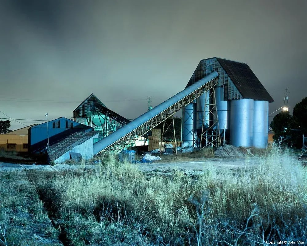 Contemporary Night Photograph "Conveyor and Silos" by John Vias: Note from the artist, John Vias: "I had to shoot this scene through the gate in a chain link fence. Notice how the metal silos seem to glow." This 11" x 14" archival color photograph is presented in