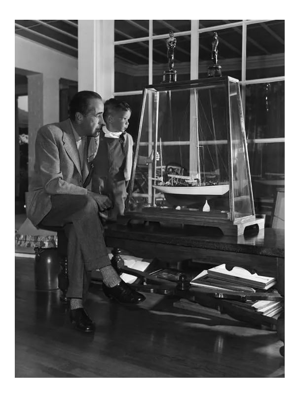 Humphrey Bogart and Son, Stephen, at Home 1952 (Photo by Sid Avery): In a quiet moment with his son, Stephen, Humphrey Bogart views a model of his yacht, the "Santana," 1952. 11" x 14" silver gelatin fiber based print signed on verso by the photographer Sid Avery. Edit