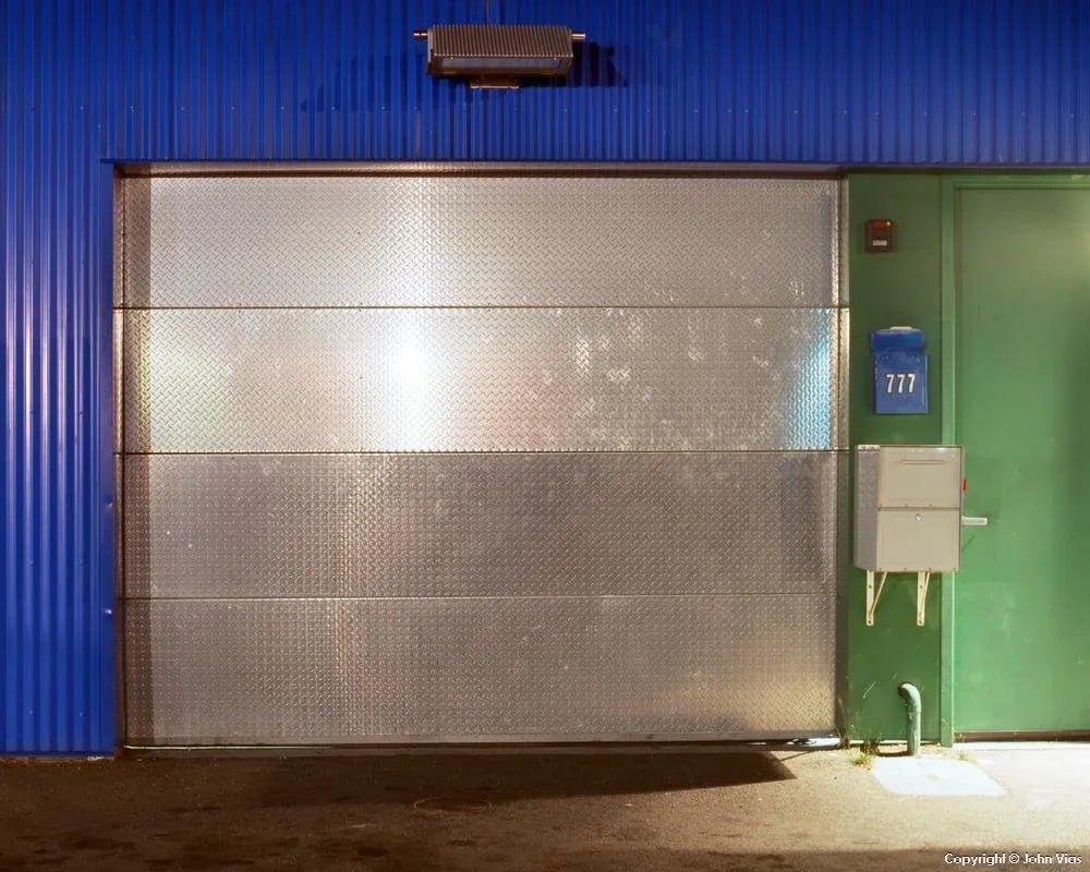 Contemporary Night Photograph "Shiny Door" by John Vias: Note from the artist, John Vias: "It is amazing how many corrugated metal buildings you can find once you start looking." This 11" x 14" archival color photograph is presented in a 16" x 20" archival