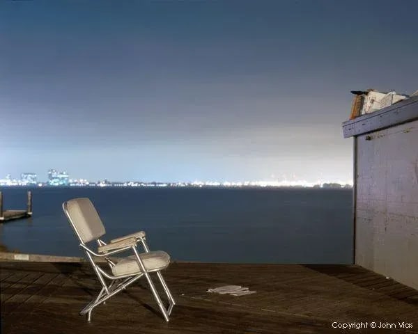 Contemporary Night Photograph "Padded Chair" by John Vias: Note from the artist, John Vias: "Why does this chair face a blank wall?" This 11" x 14" archival color photograph is presented in a 16" x 20" archival white mat. Condition: Imperfections: No