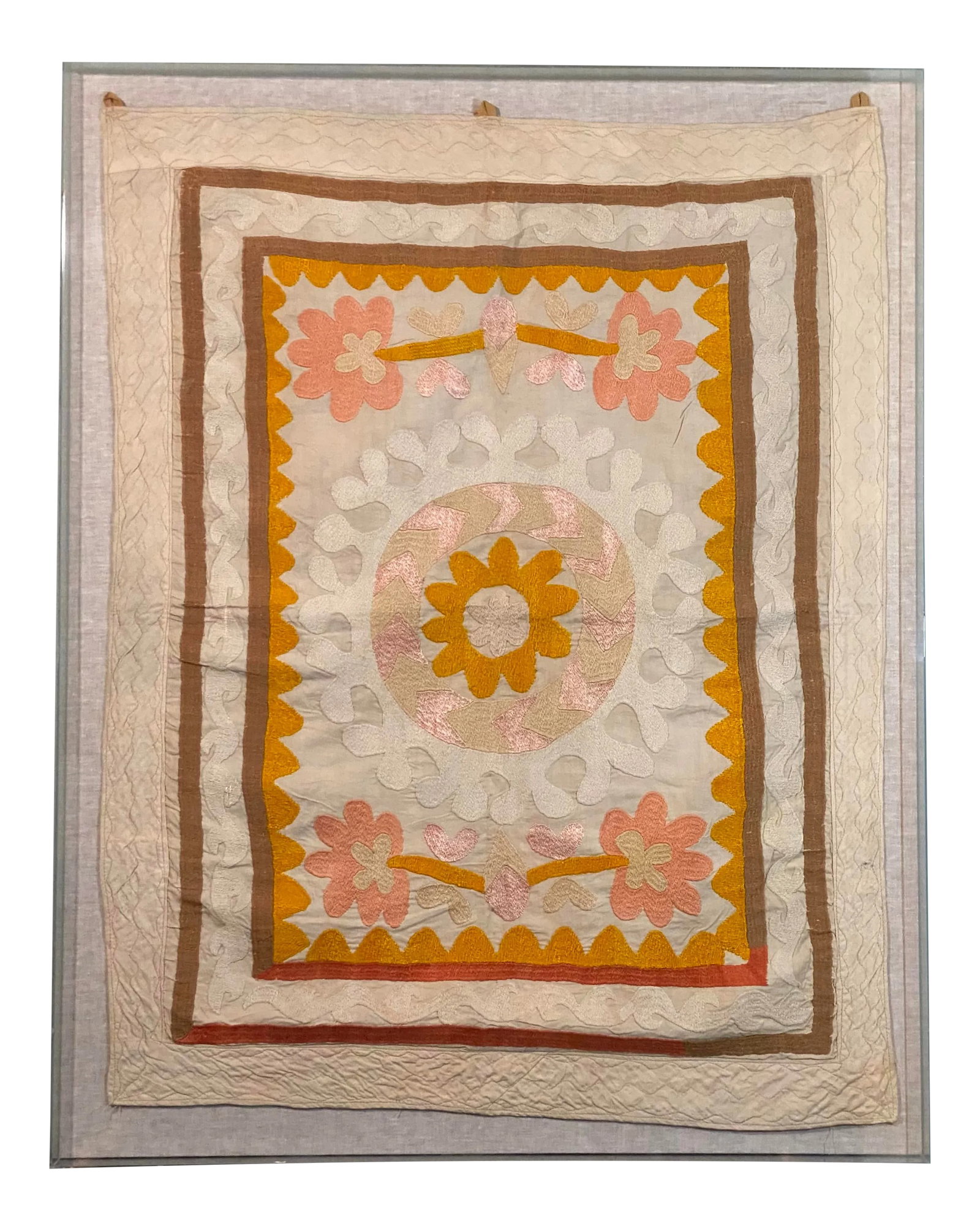 Suzani Tapestry in Acrylic Box Frame: This is a vintage Suzani tapestry from Uzbekistan. The suzani itself features a overall geometic floral design in shades of yellows, oranges, neautrals, and pale pinks or peach. The background is a
