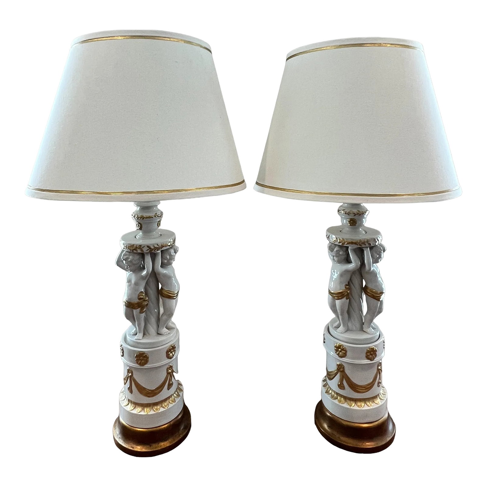 1960s Hollywood Regency Cherub-Themed Table Lamps in White Porcelain Hand-Painted With Gold + Linen: This unique Mid-Century Hollywood Regency pair of white porcelain table lamps dating to the early 1960's incorporate classic elements pulled from Italian Neoclassical and French Baroque, Rococo and Be