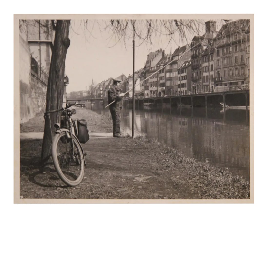 European Canal Fisherman Early Black and White Photograph: Early black and white photograph of a fisherman fishing in what appears to be a European town setting. The photograph is matted to a board to prevent bending. The photograph is not framed. Condition: