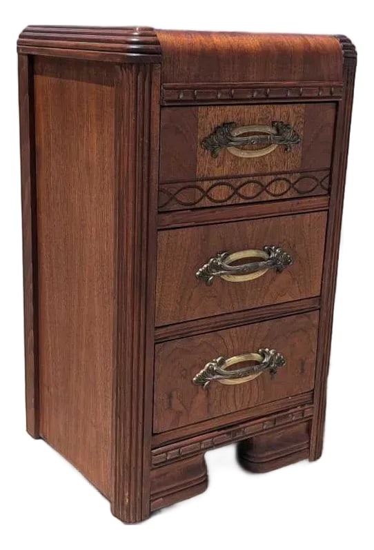 1930s Art Deco Waterfall Three-Drawer Nightstand: 1930s Art Deco Waterfall Three-Drawer Nightstand. A narrow Art Deco bedside cabinet with a rounded waterfall top, three drawers, fluted edge details, decorative banding, and ornate metal pulls. The co