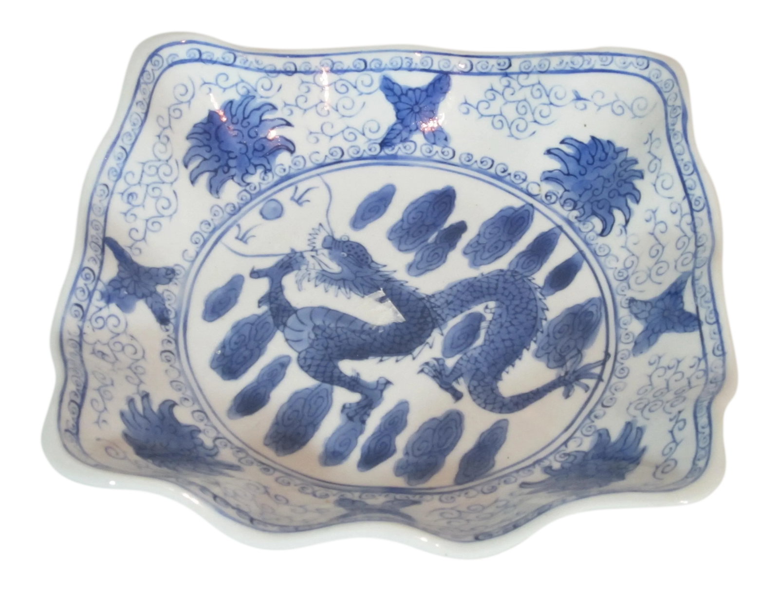 1990s Asian Porcelain Dragon Bowl: Hand painted blue and white Asian bowl decorated with a center dragon, clouds scrolls and flowers. It is square shaped with wavy sides and edges. Asian mark on the underside. Condition: Alterations: