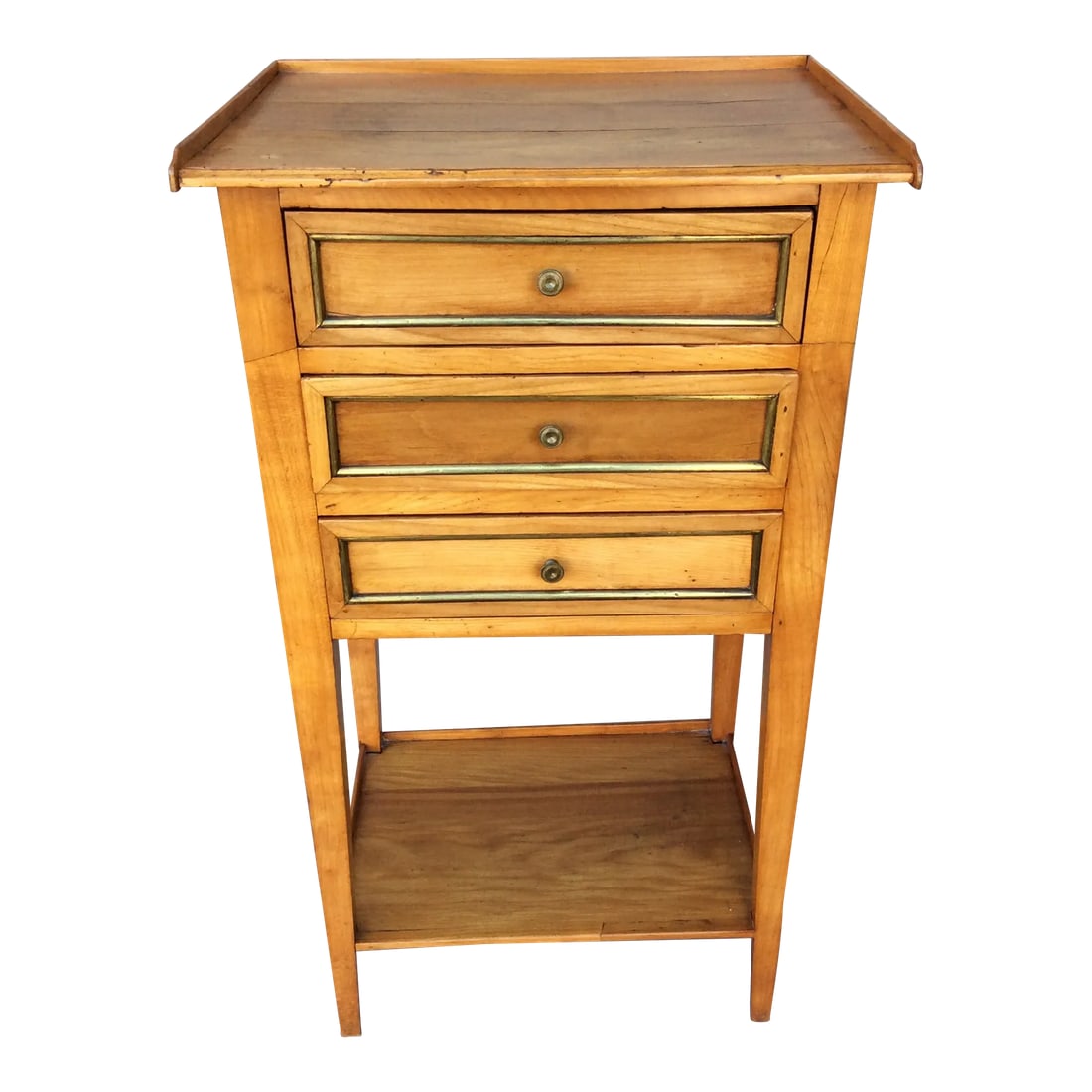 19th Century French Three Drawer Table With Brass Inlay: This 19th century French walnut side table features three drawers and a lower shelf. The drawers are embellished with small brass knobs and a brass inlay. The lower shelf provides additional storage. 