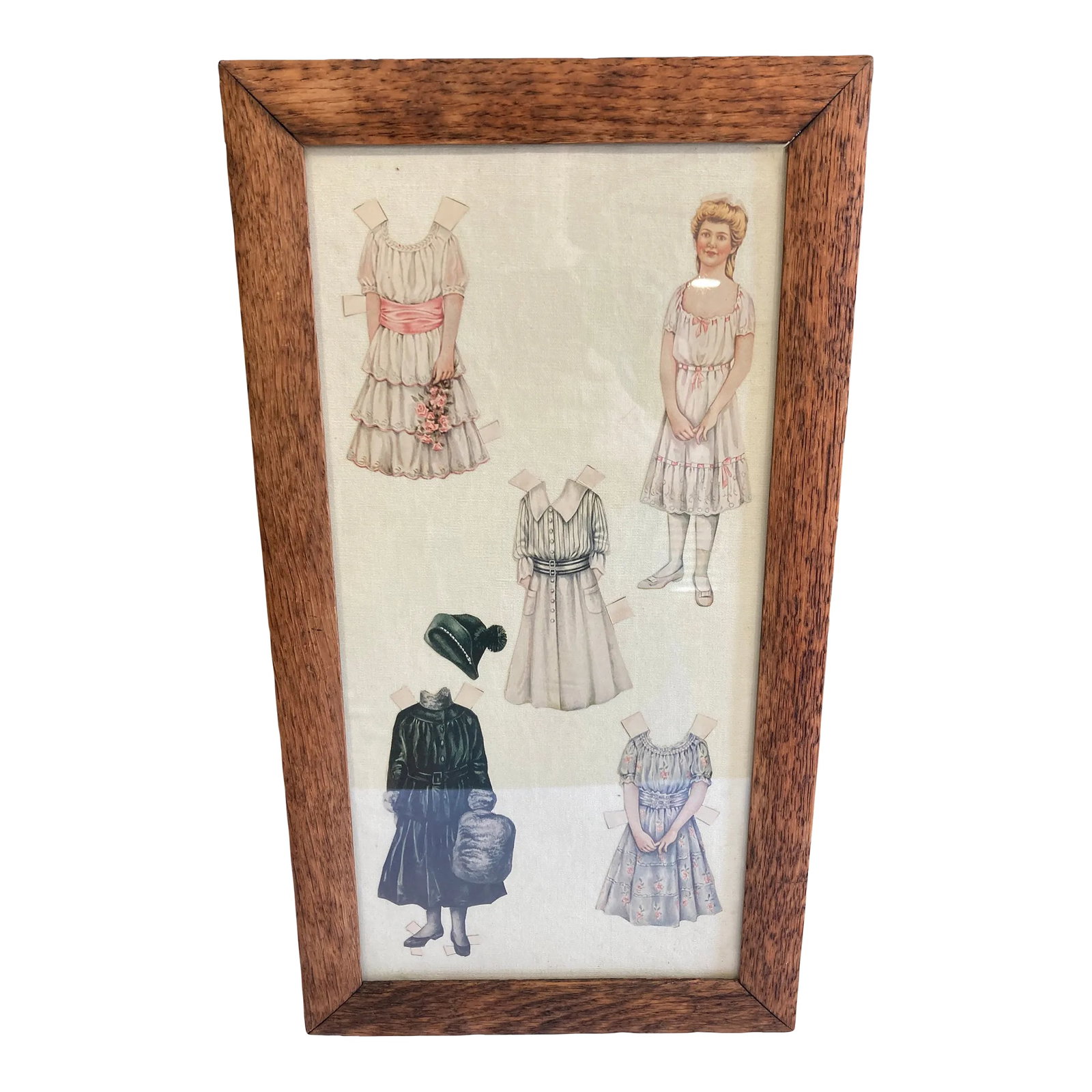 Vintage Paper Doll Clothing on Cloth Background, Framed: This framed paper dolls illustration dated 1984 showcases charming vintage-inspired figures and dresses displayed behind glass in a simple wood frame. The soft tones and nostalgic subject matter make 