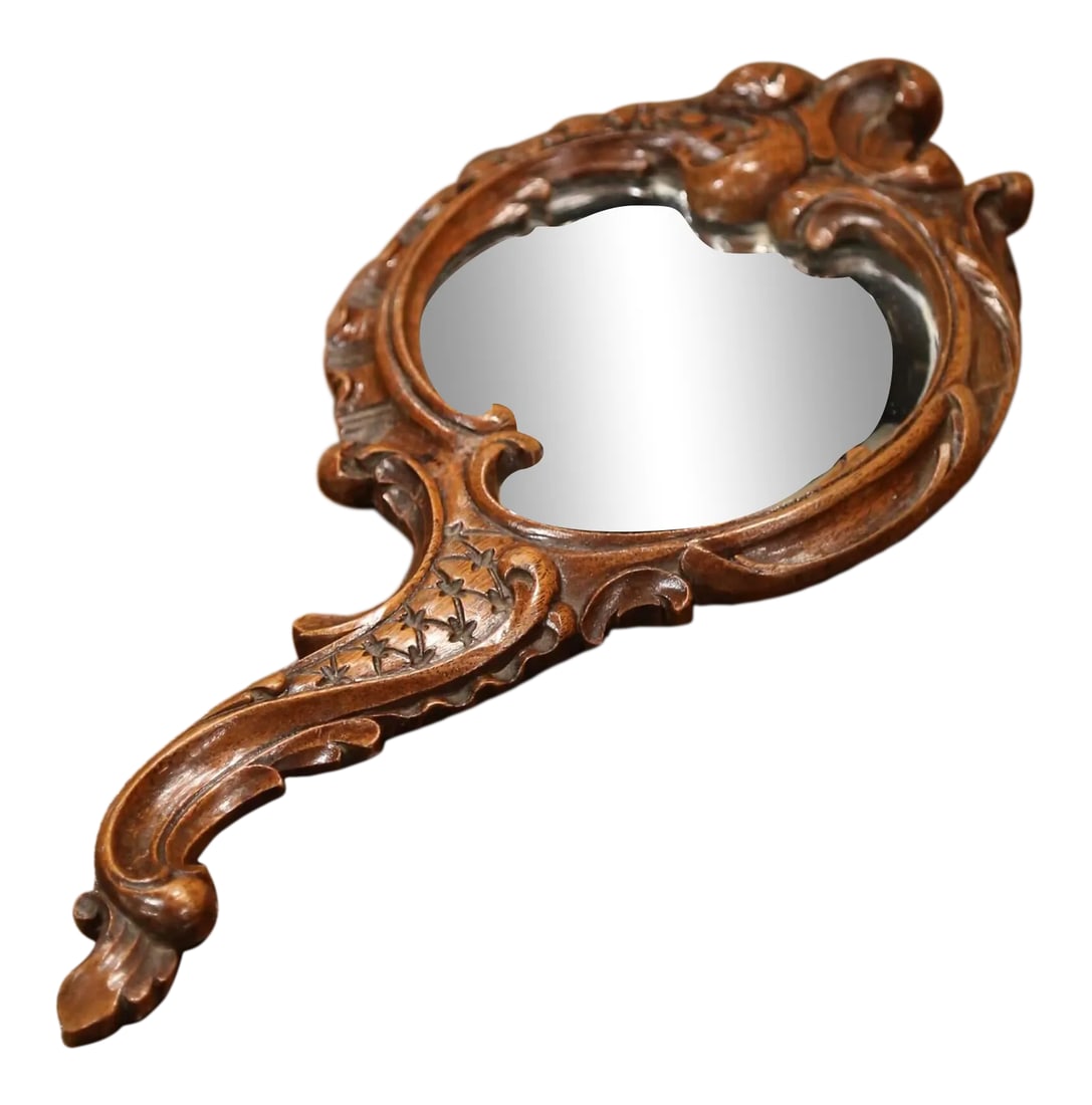 19th Century French Black Forest Carved Walnut Hand Mirror With Beveled Glass: Crafted in France circa 1870, the antique Louis XV style hand mirror with elegant serpentine handle, features hand carved floral and leaf motifs throughout the frame; the vanity mirror has the origina