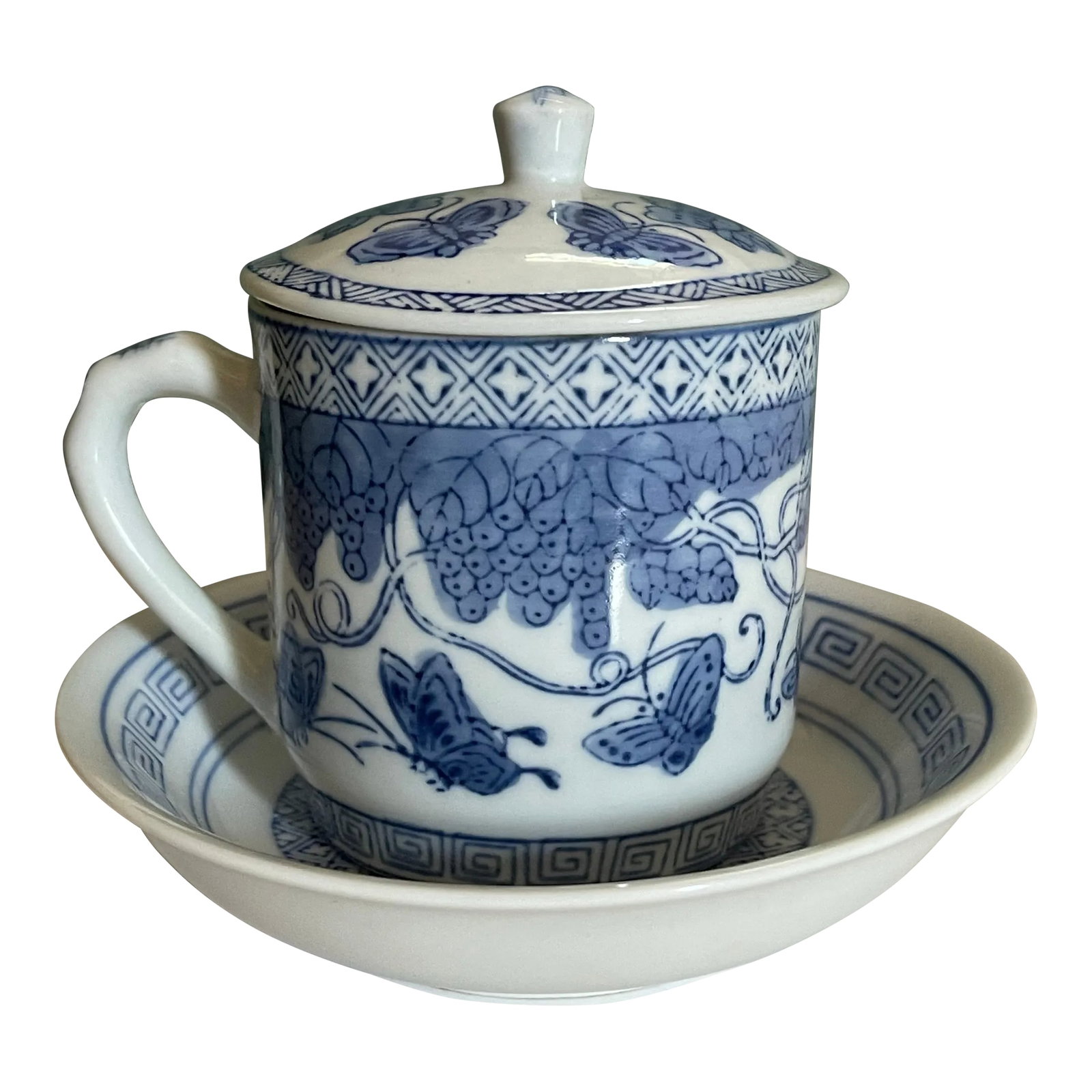 Early 20th Century Blue and White Butterfly Mug: A beautiful lidded mug with small ceramic bowl shaped saucer in a geometric pattern featuring grapes, flowers and butterflies. -Bowl measures 6 inches wide and 1 1/2 inches tall. -Mug measures 4 1/2