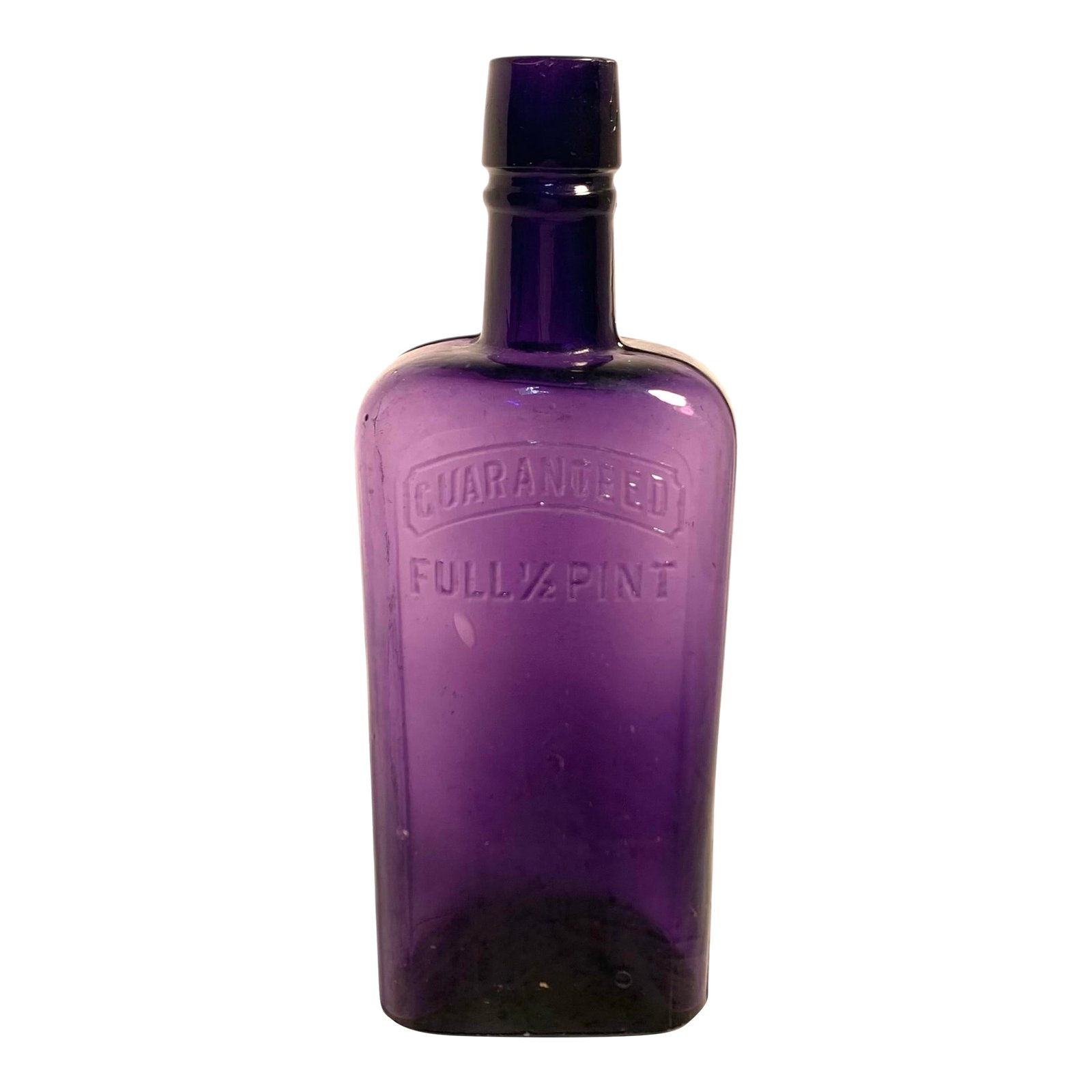 Early 20th C. Amethyst Embossed Half-Pint Whiskey Bottle: This early 20th-century whiskey bottle features bold embossed text reading "GUARANTEED FULL 1/2 PINT", a mark commonly used on pre-Prohibition liquor and medicinal containers. The bottle is made of ri