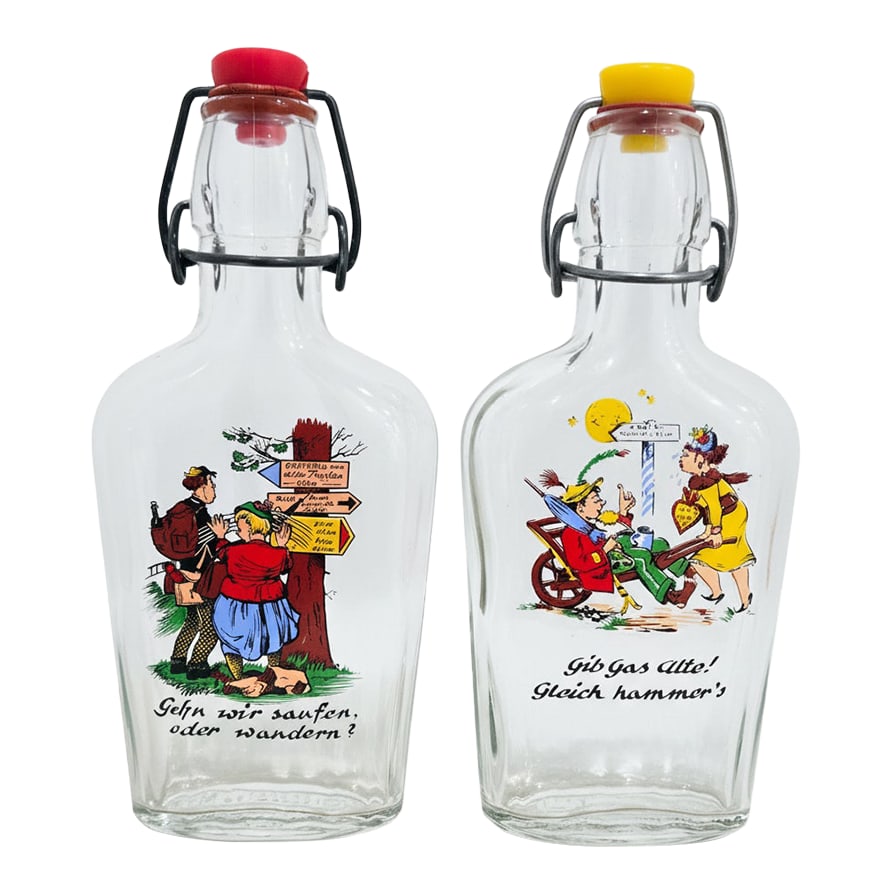 Vintage German Cartoon Swing-Top Liquor Bottles - Pair: Vintage German swing-top liquor bottles featuring colorful cartoon scenes and cheeky German phrases make a playful addition to any bar cart or display shelf. Crafted in clear glass with classic