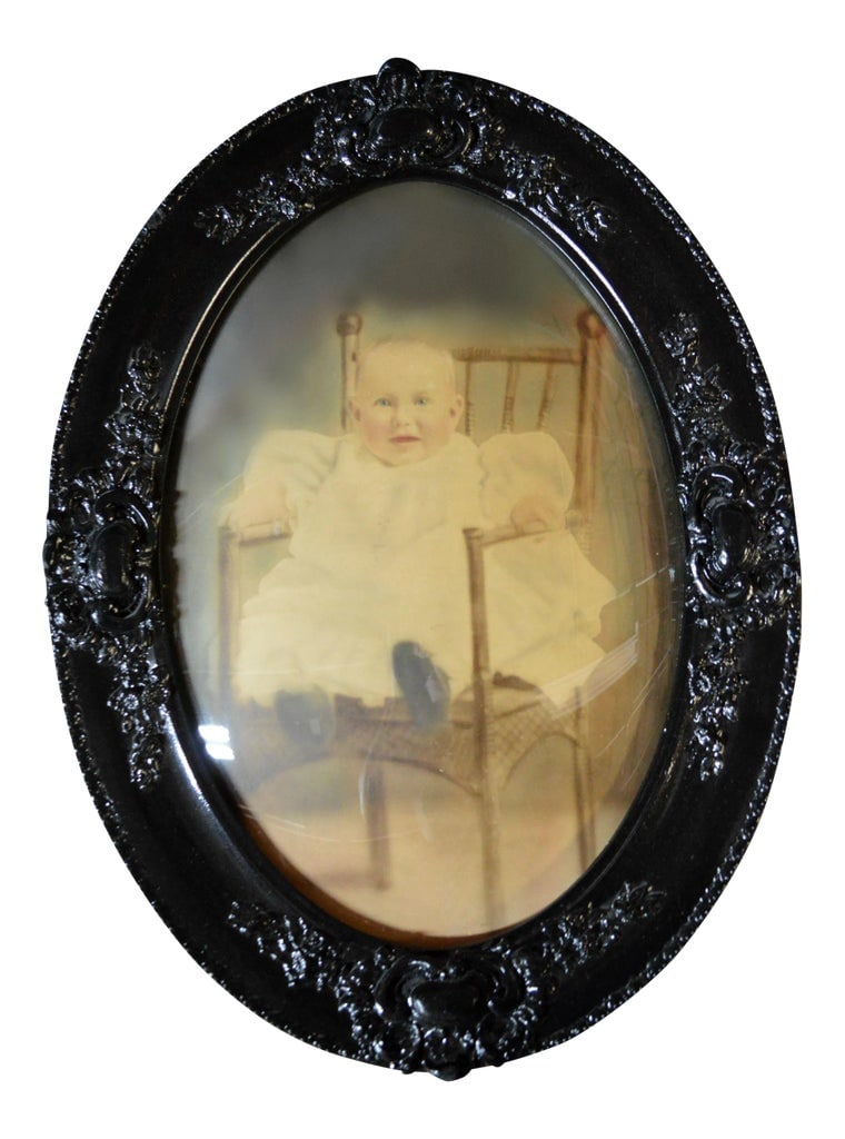 Antique Framed Baby Photograph Under Convex Glass: This is a gorgeous very old oval, manually colored, picture of a baby seated on a wicker chair. Framed in black enameled wood frame with oval bubble or convex glass. Condition: Alterations: Restored