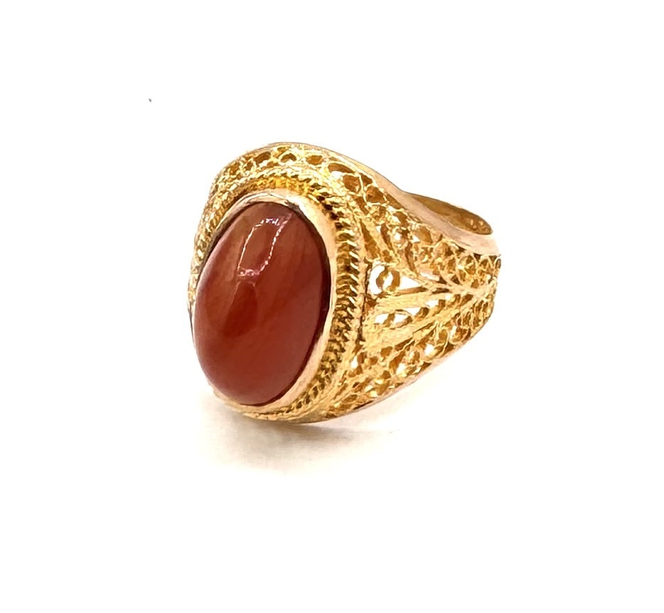 A CARNELIAN AND FILIGREE COCKTAIL RING An intricate openwork design in 18k gold: A captivating statement ring centering a vibrant oval cabochon carnelian, measuring approximately 12 x 8 mm. The gemstone displays a rich, translucent burnt-orange hue, characteristic of hi