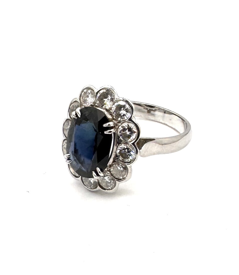 A FINE SAPPHIRE AND DIAMOND CLUSTER RING A classic "Royal" silhouette of exceptional proportions in: An elegant and high-impact sapphire dress ring, masterfully crafted in 18k white gold (750/1000). The design is centered upon a magnificent oval-cut blue sapphire, measuring approximately 11 x 9 mm. T