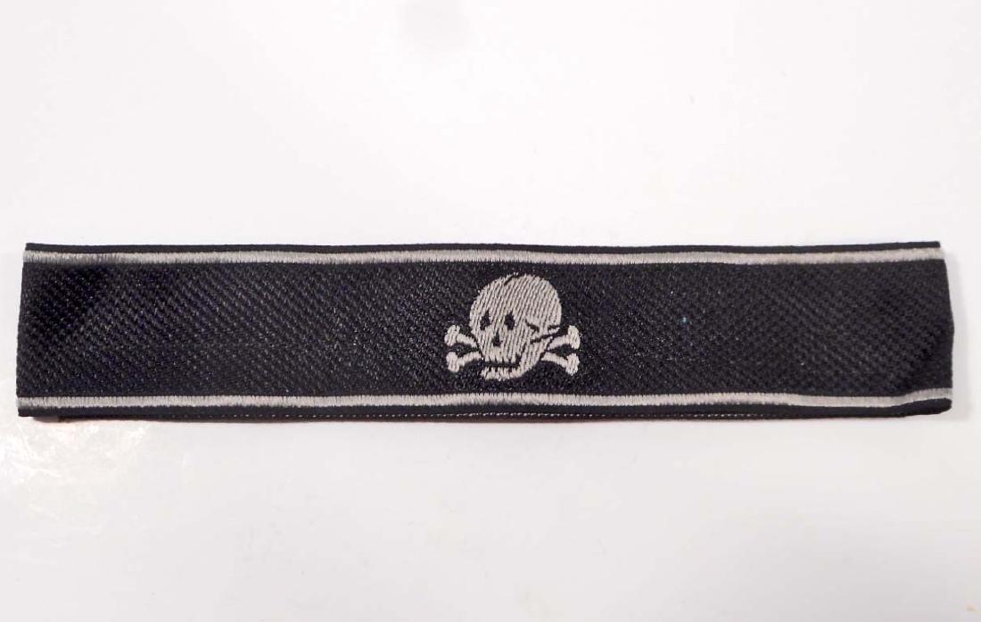 GERMAN NAZI WAFFEN SS TOTENKOPFEM CUFF TITLE: Measures 20 1/8" wide by 1 1/4" tall. The front has a skull in the center. Has a salt and pepper back. Machine embroidered bevo woven cloth construction. This was to have been worn on the lower left s