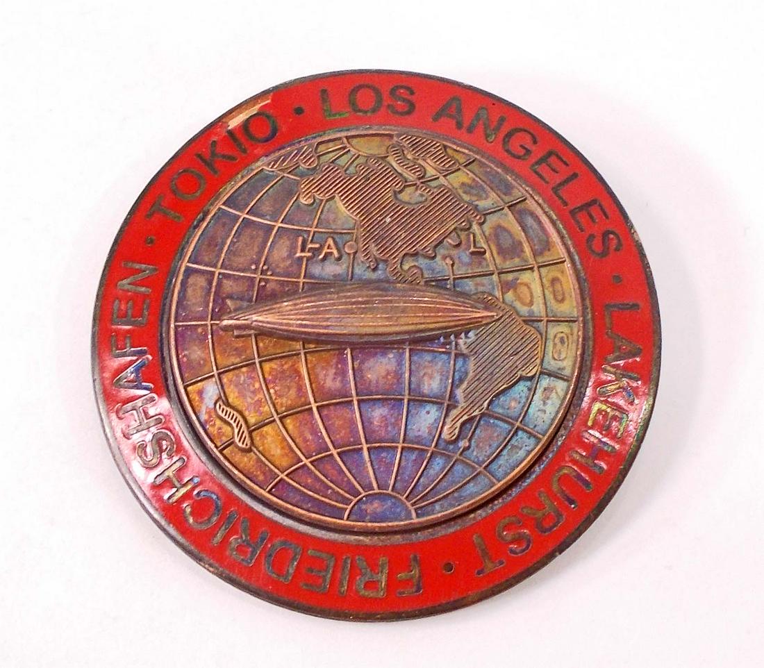 GERMAN NAZI GRAF ZEPPELIN TOKIO LOS ANGELES LAKEHURST: Measures 1 3/4" in diameter. The front reads 'Tokio - Los Angeles - Lakehurst Friedrichshafen'. Shows the Graf Zeppelin flying over the world continent. The reverse is maker marked 'FLL'. Has a thin h