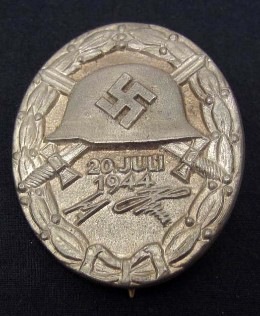 GERMAN NAZI SILVER WOUND BADGE: Thin vertical pinback. Maker arked C E Junker Berlin SW 68. Front shows a German helmet w/ a pair of crossed swords behind. Below reads 20 Juli 1944 Adolf Hitler. Has an oak leaf wreath around the edg