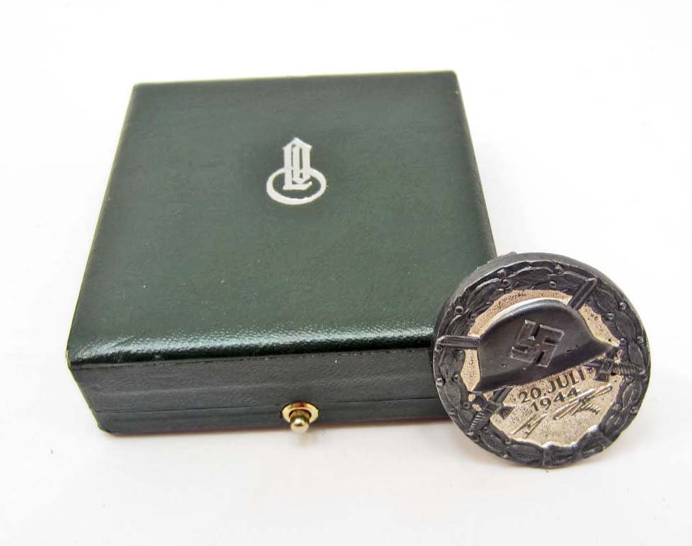 CASED GERMAN NAZI JULY 20TH BLACK WOUND BADGE: Has a wide vertical pinback. Maker marked 2 800. Front shows a german helmet w/ a swastika in the center w/ a pair of crossed swords in the back ground. Below reads 20 Juli Adolf Hitler. Blued nickel 