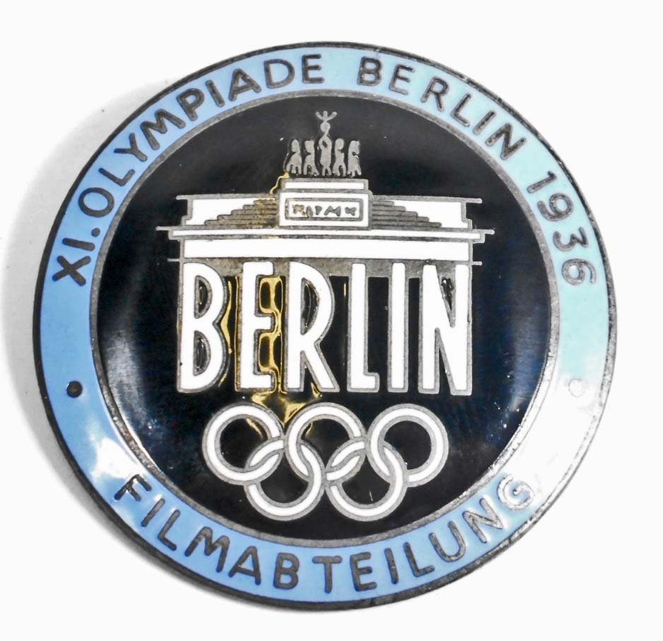 GERMAN NAZI BERLIN SUMMER OLYMPICS FILM MAKERS BADGE: Measures 1 9/16" in diameter. The reverse side is maker marked 'Rob Neff Berlin W 57'. Has a thin horizontal pin back. One piece nickel construction with black, white and blue enamel.
