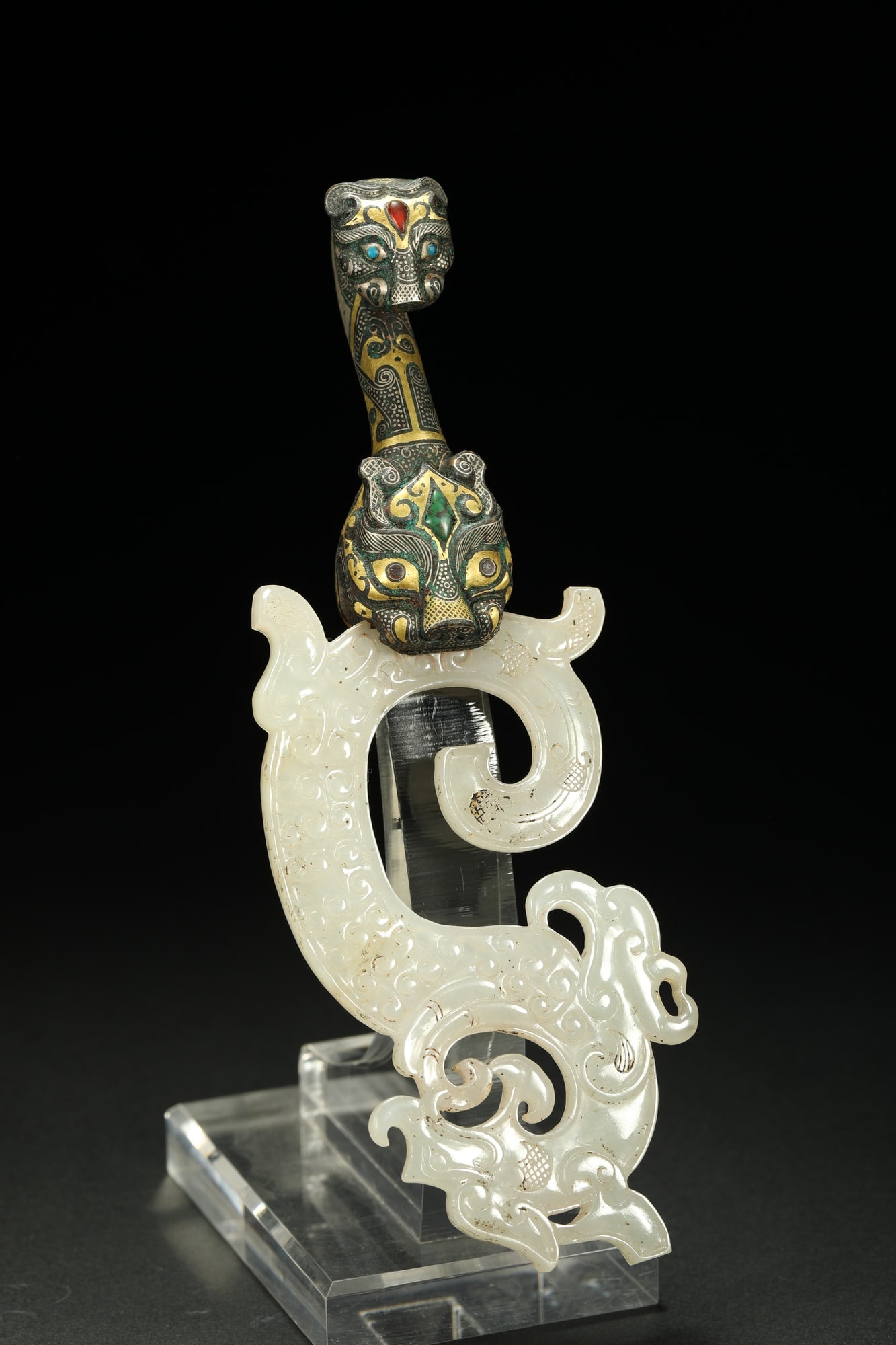 BRONZE GILT-SILVER JADE BELT HOOK: Dimensions: 7 1/4 x 2 3/8 in; weight 206 g This bronze gilt-silver jade belt hook presents an elegant fusion of jade carving and intricate metalwork decoration. The curved hook form is enriched by